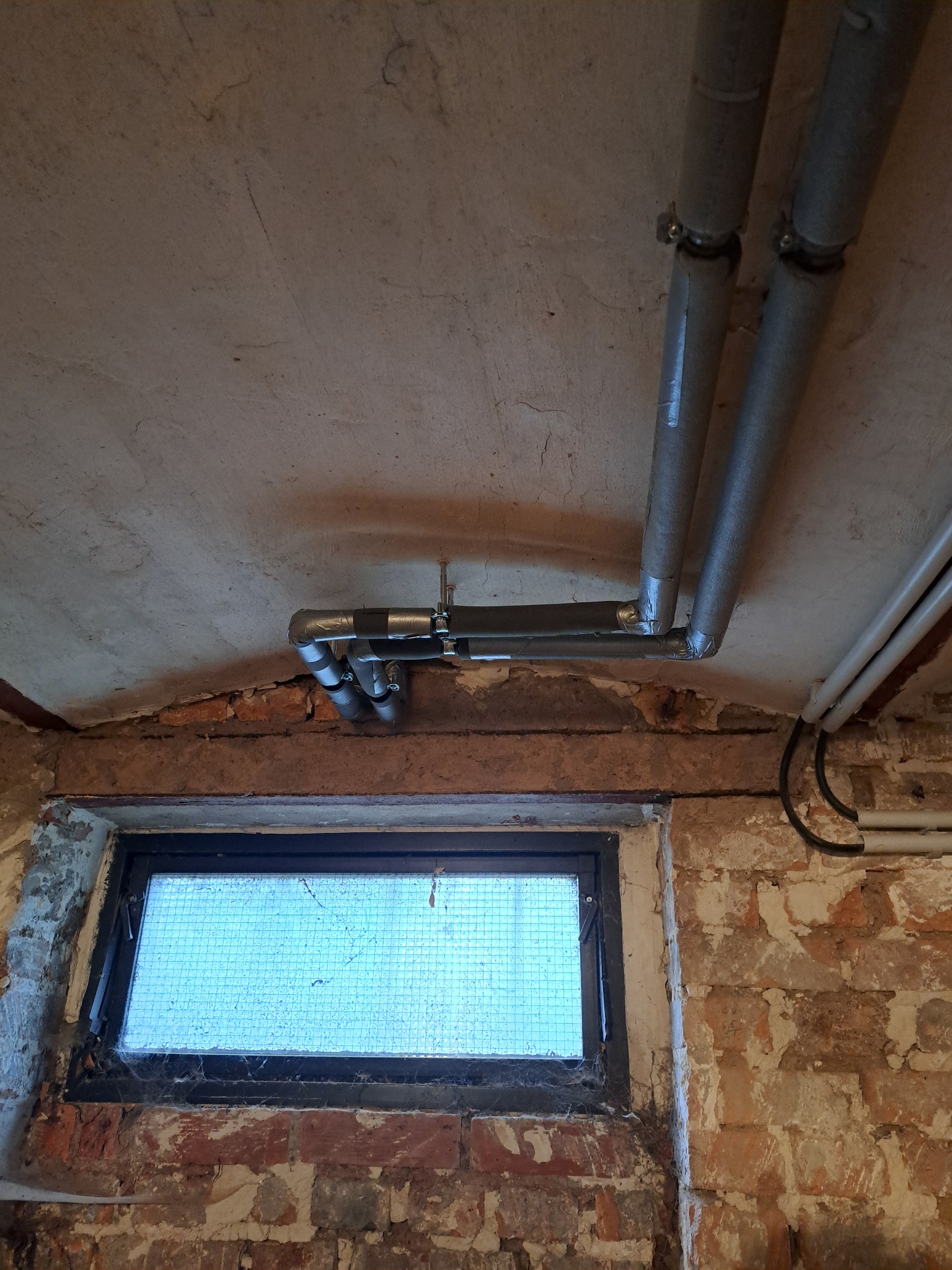 Szare, zaizolowane rury grzewcze przymocowane do sufitu w piwnicy z małym oknem i odsłoniętą cegłą. Widoczne ślady napraw i izolacji.