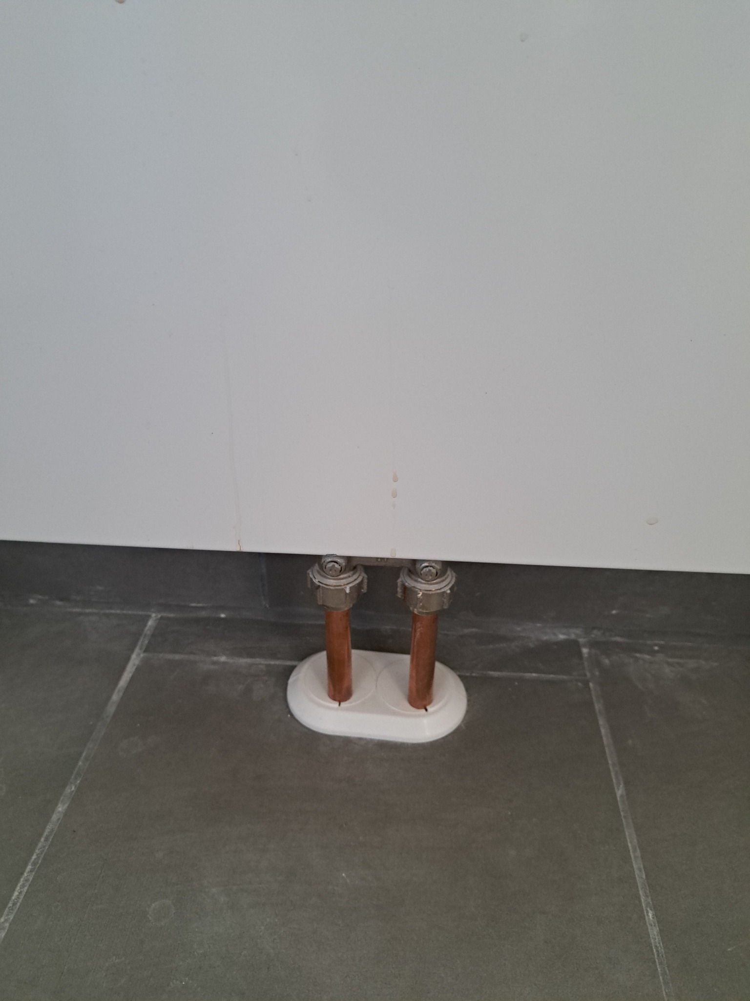 Miedziane rury grzewcze wystające z podłogi, zabezpieczone białą osłoną, widoczne na tle szarej płytki podłogowej i białej ściany.