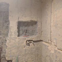 Usługi budowlane Daniel Suchecki - Surowa ściana w trakcie prac wykończeniowych z widocznymi instalacjami i wnęką. Fragment drewnianej listwy w dolnej części kadru. Ściana pokryta warstwą szpachli.