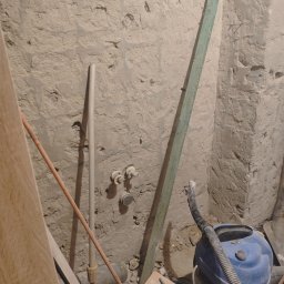 Usługi budowlane Daniel Suchecki - Surowa ściana z instalacją wodną w trakcie prac. Widoczne rury, listwa, odkurzacz i dokumentacja techniczna. Przygotowanie do wykończenia wnętrza.