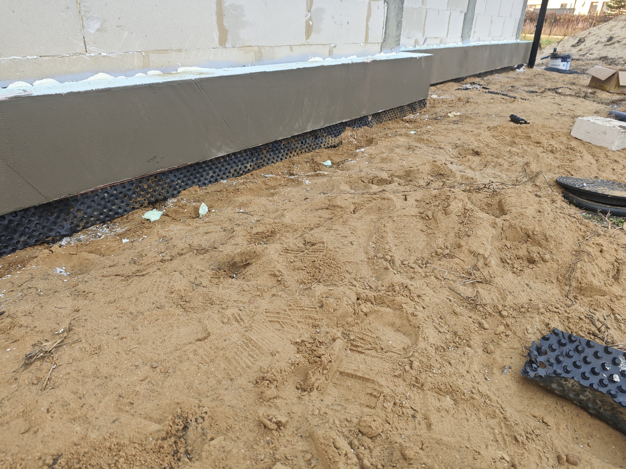 Ocieplenie fundamentów budynku z widoczną folią kubełkową i warstwą izolacyjną. Prace ziemne w toku, piasek wokół. Detal budowlany.