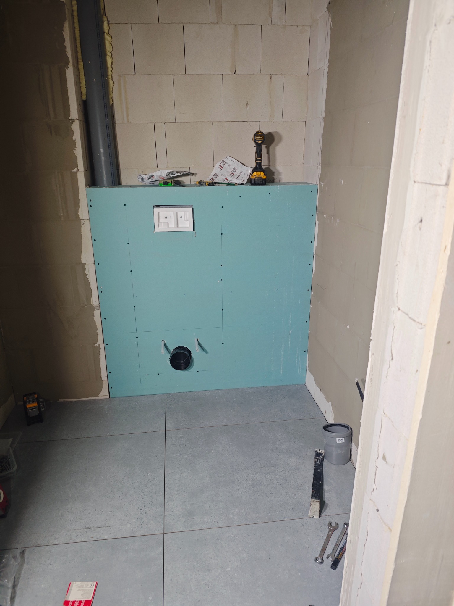 Niebieska płyta GK z otworem na WC i przyciskiem spłukiwania, zamontowana w surowej łazience z szarymi płytkami podłogowymi. Widoczne narzędzia i elementy instalacji.