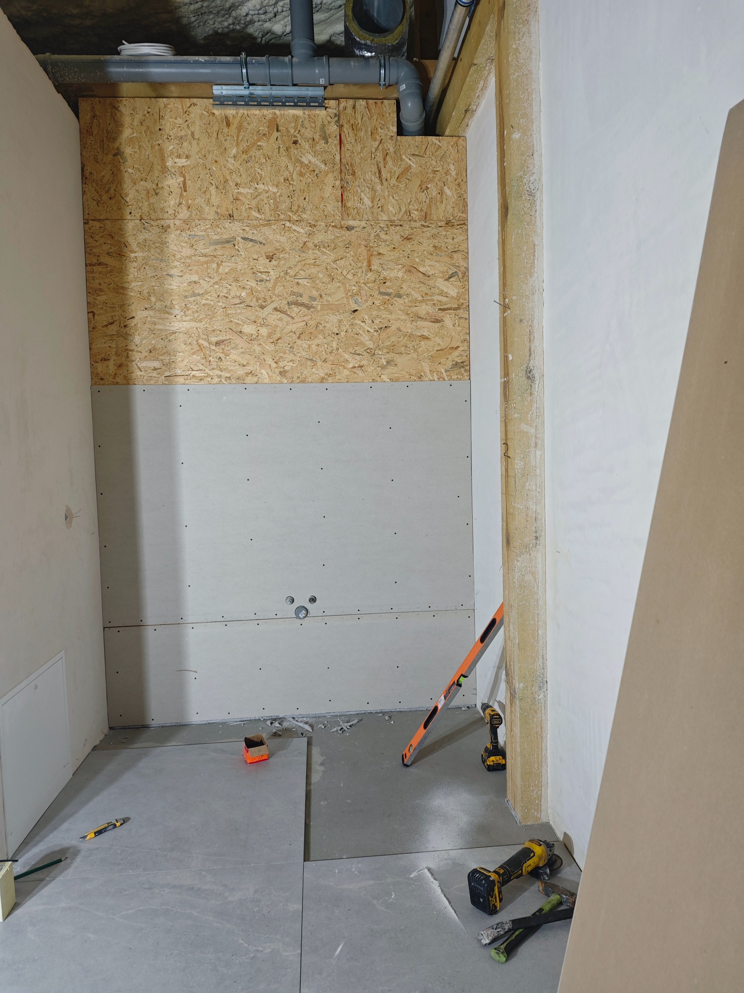 Łazienka w trakcie remontu: płyty OSB, gips-karton, częściowo ułożone szare płytki podłogowe, poziomica i narzędzia. Widoczne rury pod sufitem.