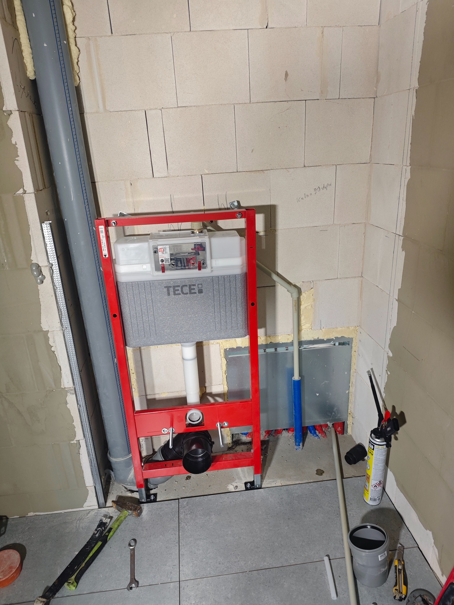 Montaż stelaża podtynkowego TECE w surowej łazience z widocznymi instalacjami i narzędziami. Ściany z betonu komórkowego, szare płytki podłogowe.