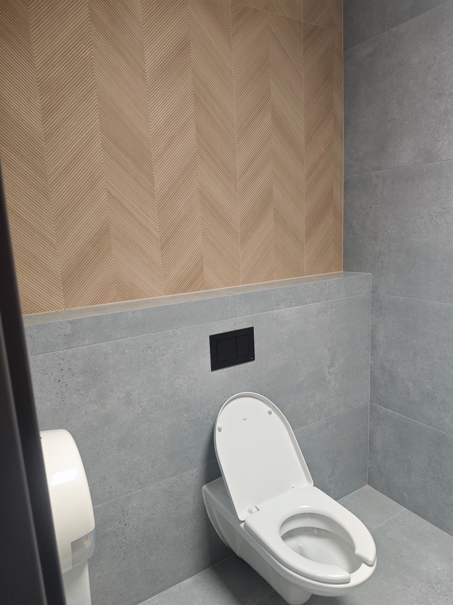 Nowoczesna łazienka z toaletą wiszącą, szarą glazurą i drewnopodobną ścianą w jodełkę. Minimalistyczny design, czarny przycisk spłukiwania, dozownik papieru.