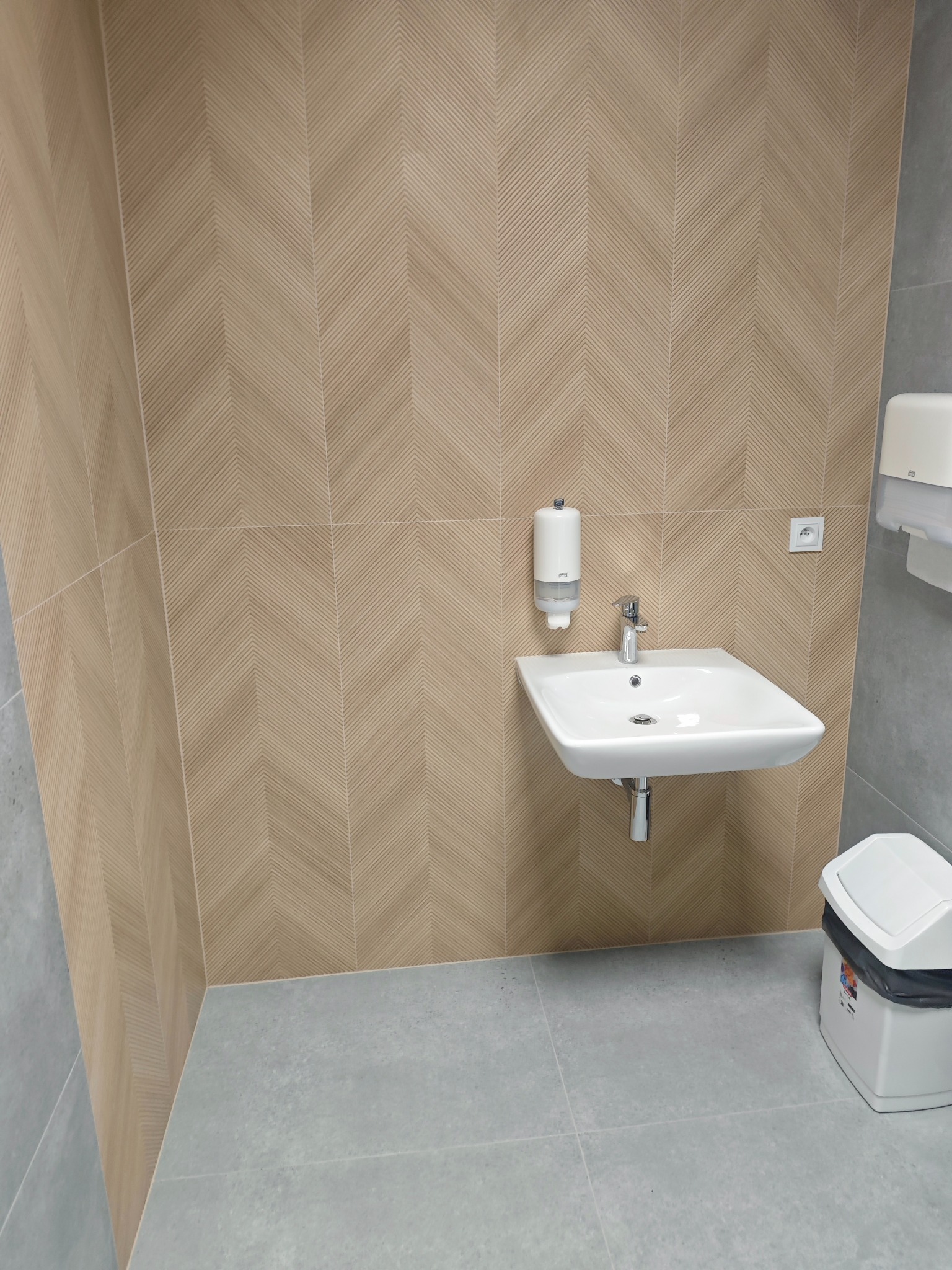 Nowoczesna łazienka z umywalką i beżowymi płytkami ułożonymi w jodełkę na ścianie. Szare płytki podłogowe i biały kosz na śmieci. Minimalistyczny design.