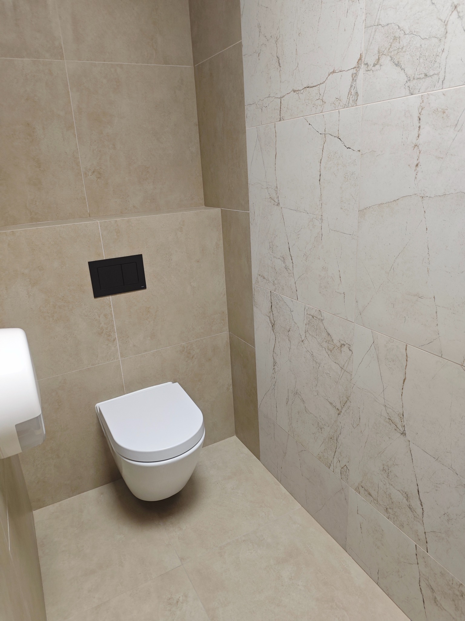 Nowoczesna toaleta podwieszana z czarnym przyciskiem spłukującym, otoczona beżowymi i marmurkowymi płytkami w łazience. Minimalistyczny design.