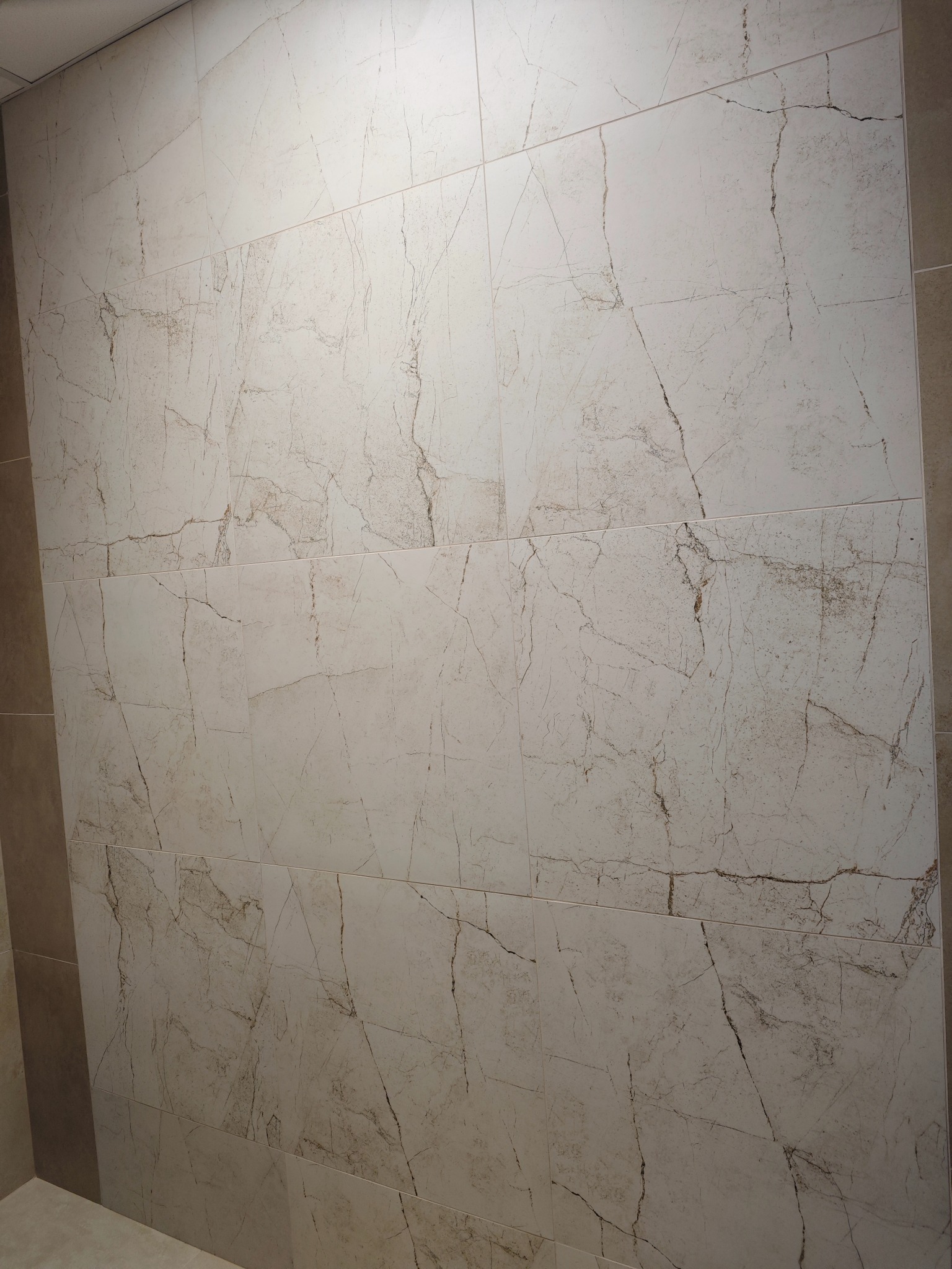 Ściana wyłożona jasnymi, marmurkowymi płytkami w łazience. Widoczne fugi i fragmenty sąsiednich ścian w odcieniach beżu. Minimalistyczny design.