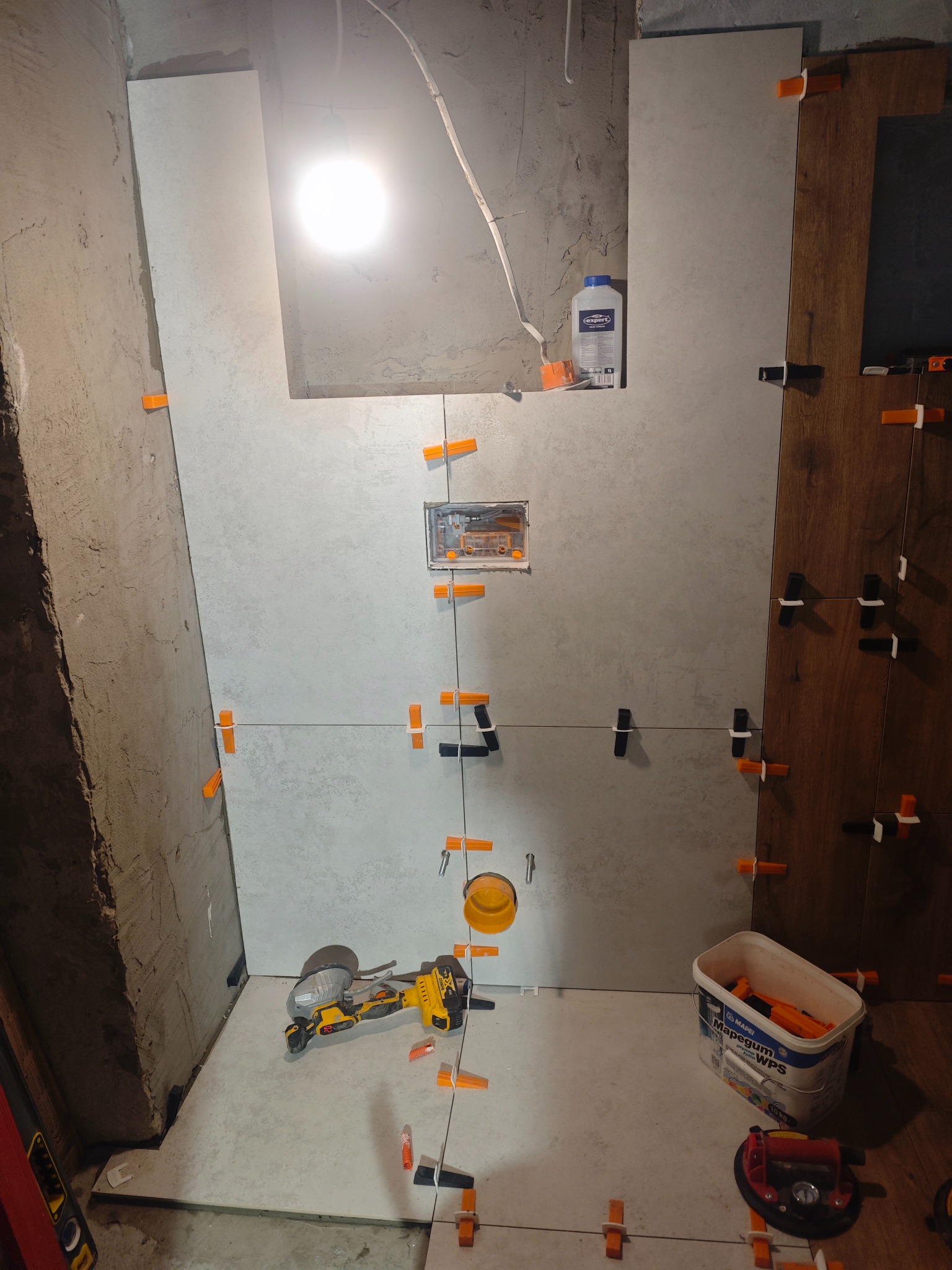 Układanie płytek w łazience z użyciem poziomowania. Widoczne narzędzia, klej i fragment drewnopodobnej okładziny. Oświetlenie halogenowe.