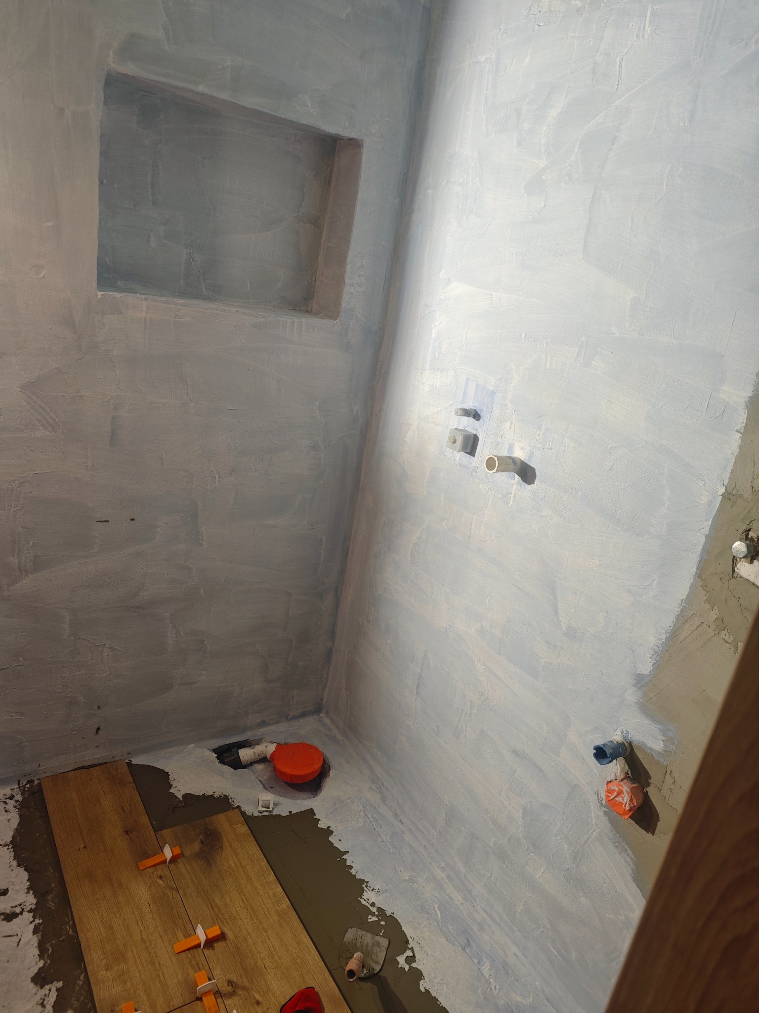 Wnętrze łazienki w trakcie prac. Ściany po gruntowaniu, częściowo ułożone płytki imitujące drewno z krzyżykami dystansowymi. Widoczna wnęka i instalacja wodna.