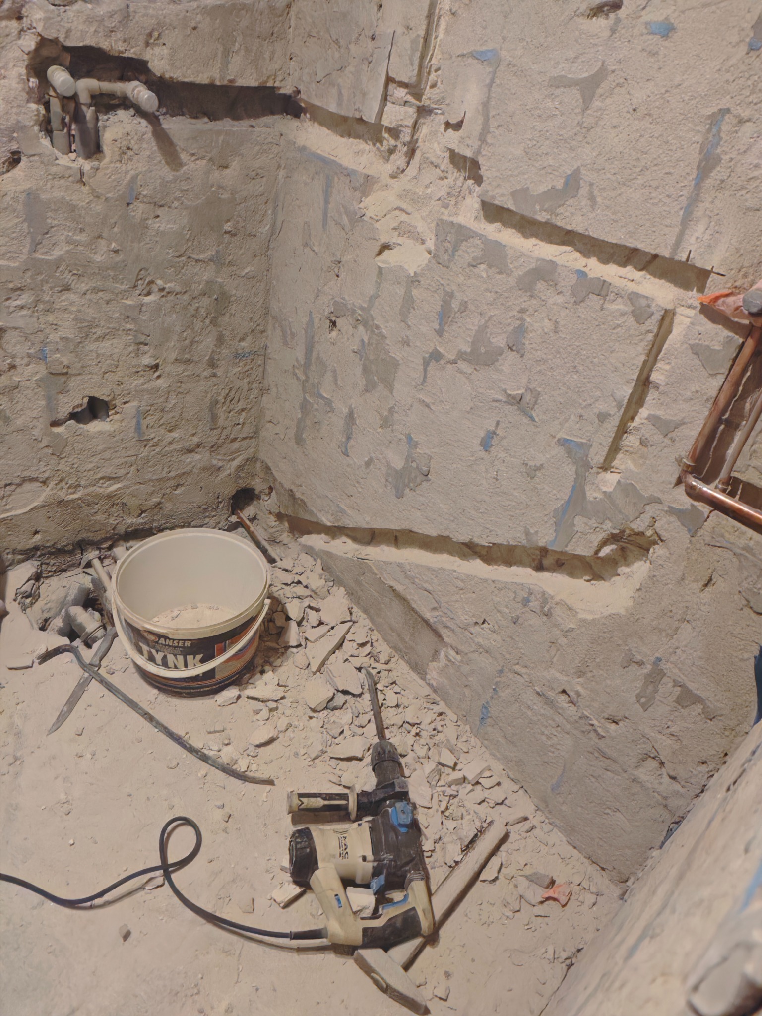 Kucie ścian w łazience pod instalacje. Widoczny młot udarowy, wiadro z zaprawą i fragment instalacji. Ściany w trakcie prac przygotowawczych.
