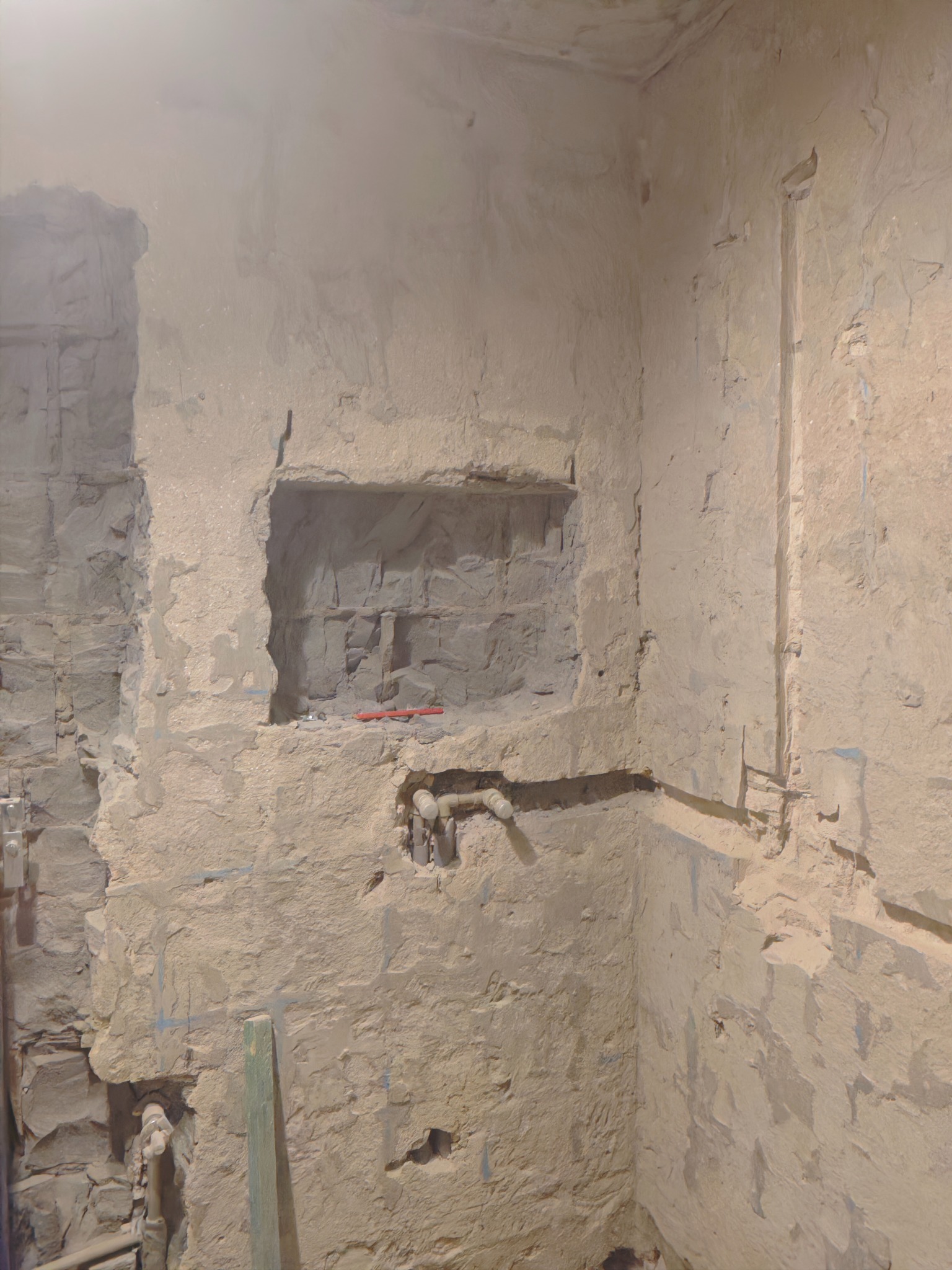 Surowa ściana w trakcie prac wykończeniowych z widocznymi instalacjami i wnęką. Fragment drewnianej listwy w dolnej części kadru. Ściana pokryta warstwą szpachli.