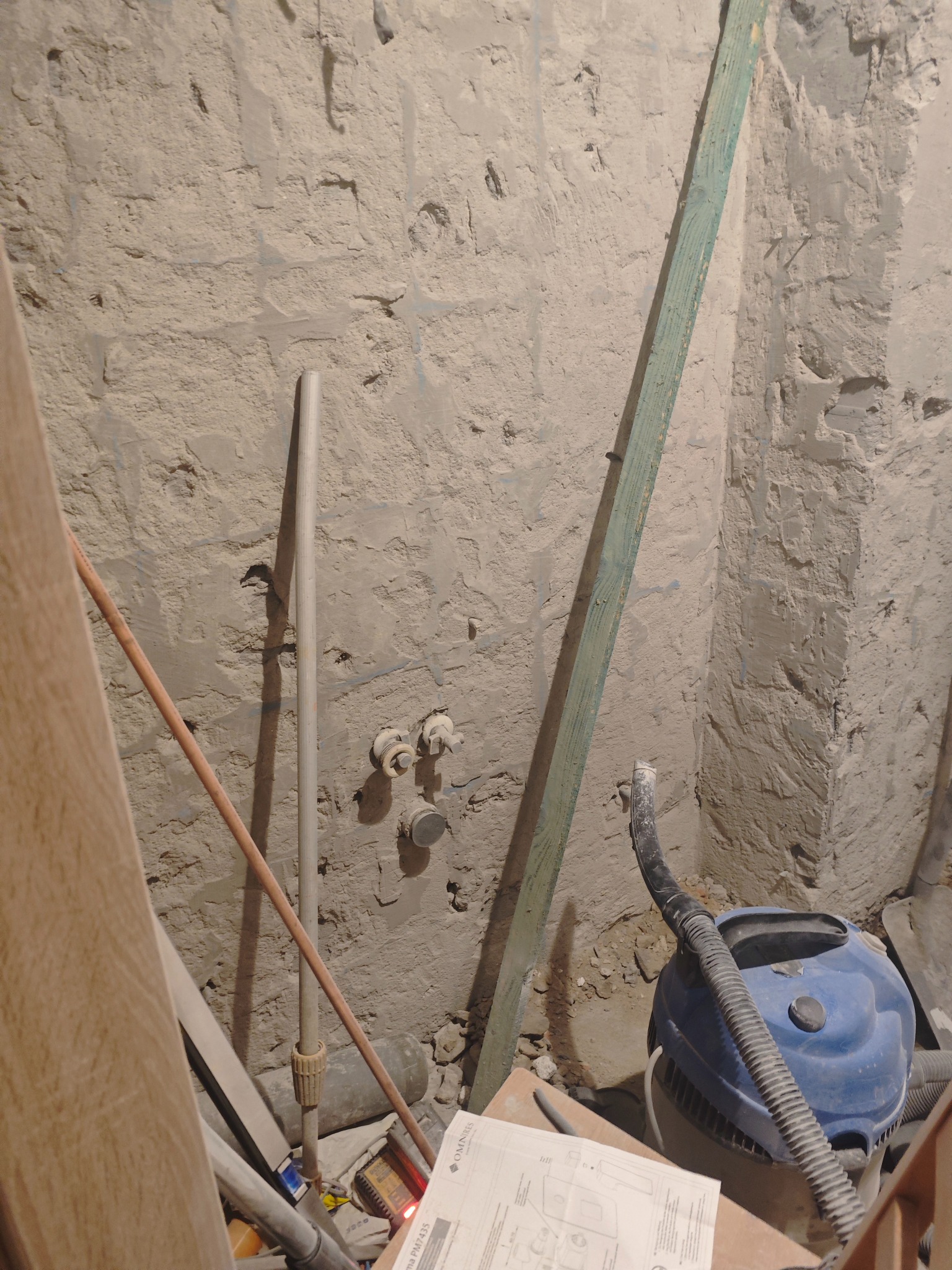 Surowa ściana z instalacją wodną w trakcie prac. Widoczne rury, listwa, odkurzacz i dokumentacja techniczna. Przygotowanie do wykończenia wnętrza.