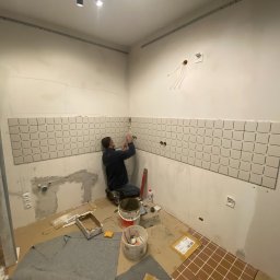Mirosław Zdeb - Układanie białych, kwadratowych płytek na ścianie w łazience. Widoczny glazurnik w trakcie pracy, narzędzia i materiały budowlane. Podłoga wyłożona brązowymi płytkami.