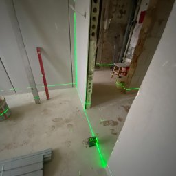 Mirosław Zdeb - Wnętrze w trakcie prac wykończeniowych z użyciem lasera krzyżowego, poziomicy i profili. Widoczne ślady stóp na podłodze i surowe wykończenie w tle.