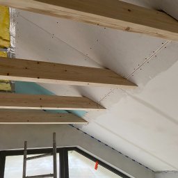 Mirosław Zdeb - Skośny sufit poddasza w trakcie wykańczania: drewniane belki, płyty gipsowe, izolacja, okno z folią ochronną, drabina. Widok z dołu, etap prac wykończeniowych.