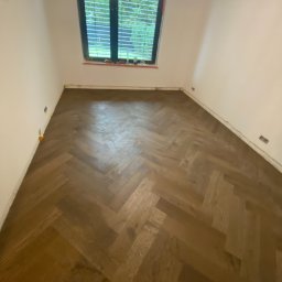 Mirosław Zdeb - Ukończona podłoga z parkietu ułożonego w jodełkę w pustym pokoju z oknem. Widoczne listwy przypodłogowe i żółte kliny dystansowe.