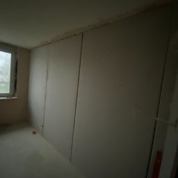 Mirosław Zdeb - Wnętrze w trakcie prac wykończeniowych: ściany pokryte płytami gipsowo-kartonowymi, widoczne łączenia. Okno z widokiem na zewnątrz. Na podłodze leży pomarańczowy klocek.