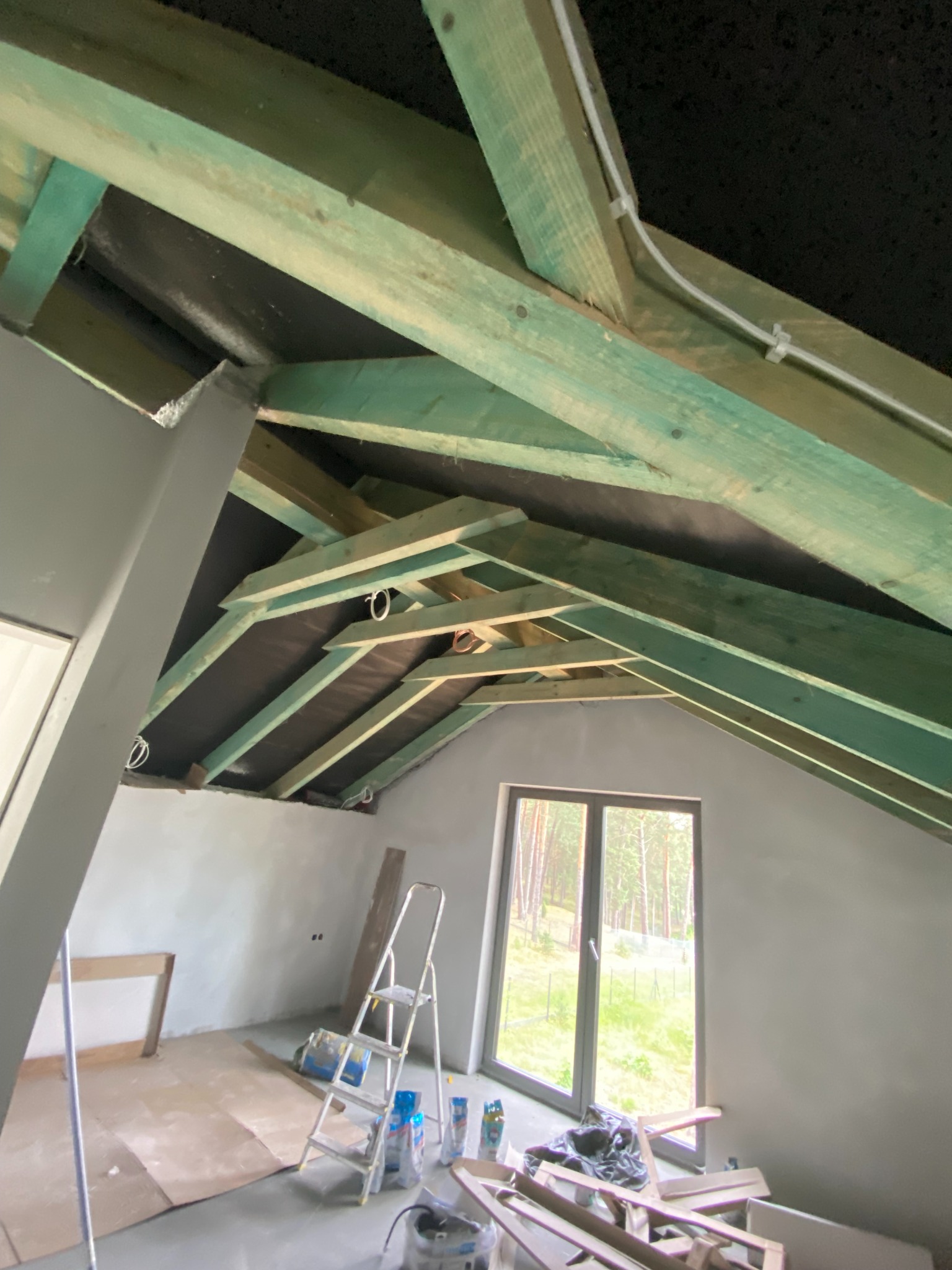 Poddasze w trakcie adaptacji: widoczne zielone belki konstrukcyjne, czarna membrana, okno z widokiem na las, drabina i materiały budowlane. Surowy stan wykończenia.