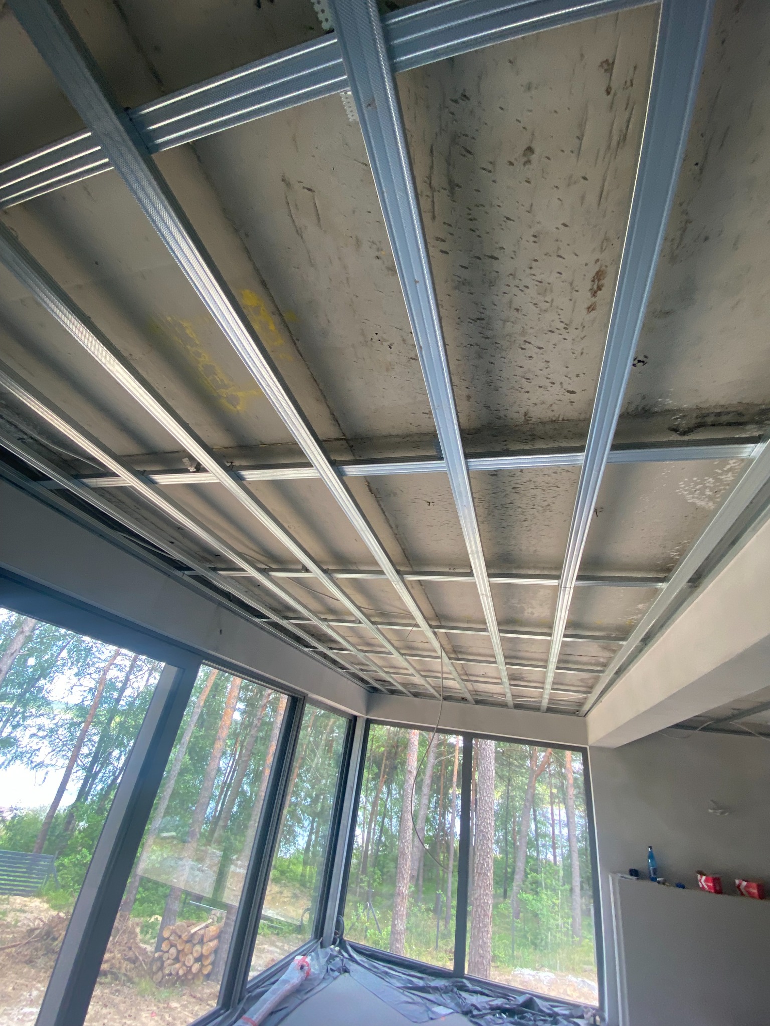 Montaż metalowego stelażu sufitu podwieszanego w jasnym pomieszczeniu z dużymi oknami, widok na las. Etap prac wykończeniowych.