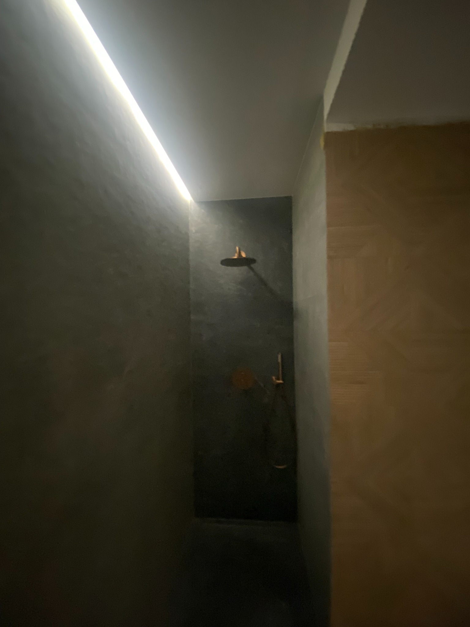 Nowoczesna kabina prysznicowa z szarą, teksturowaną ścianą, oświetleniem LED i złotą armaturą. Minimalistyczny design z widoczną fakturą ściany.