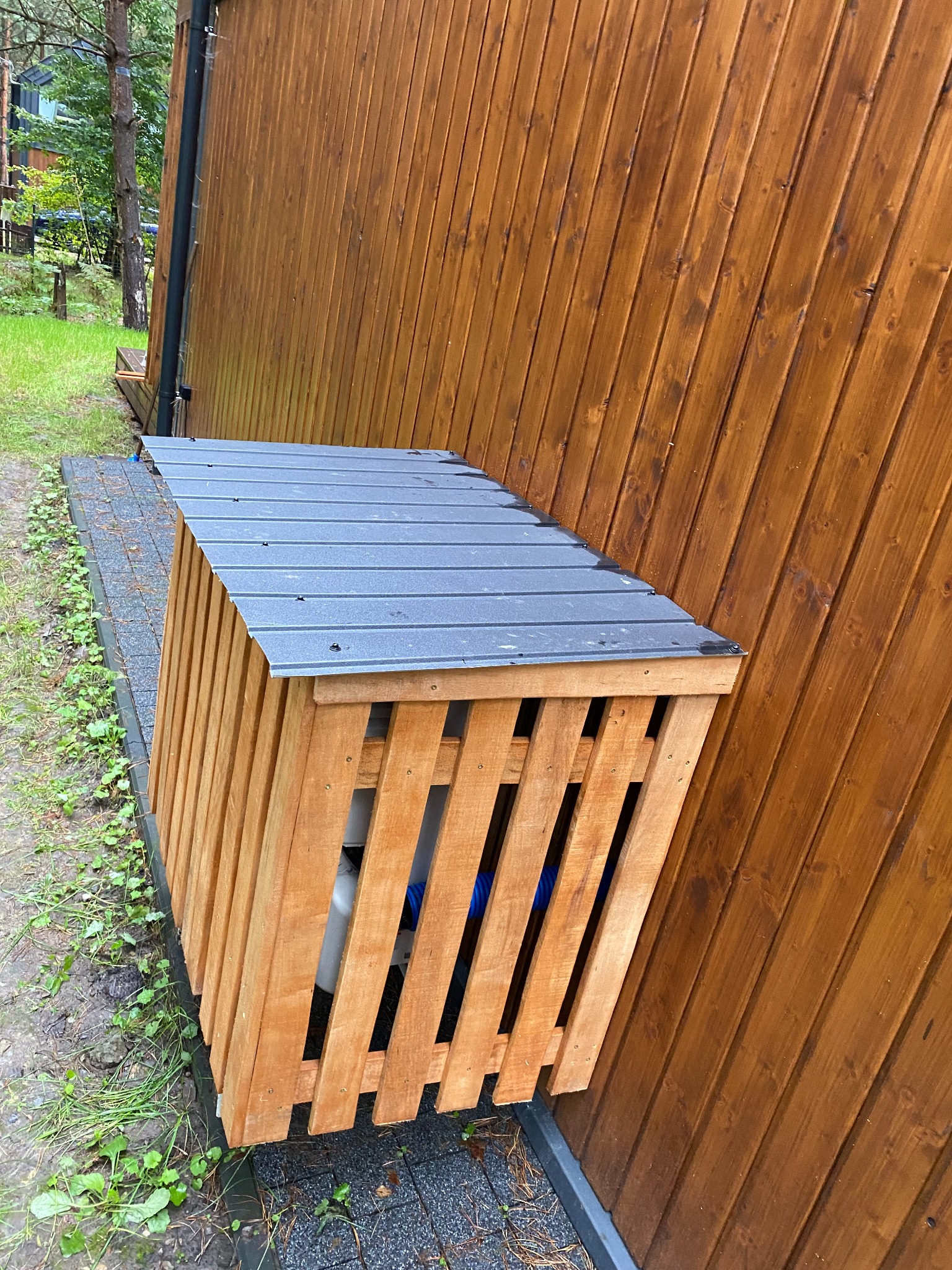 Drewniana obudowa na zewnątrz budynku, z szarą, metalową pokrywą, przylegająca do ściany z desek. Widoczne elementy instalacji wewnątrz obudowy.