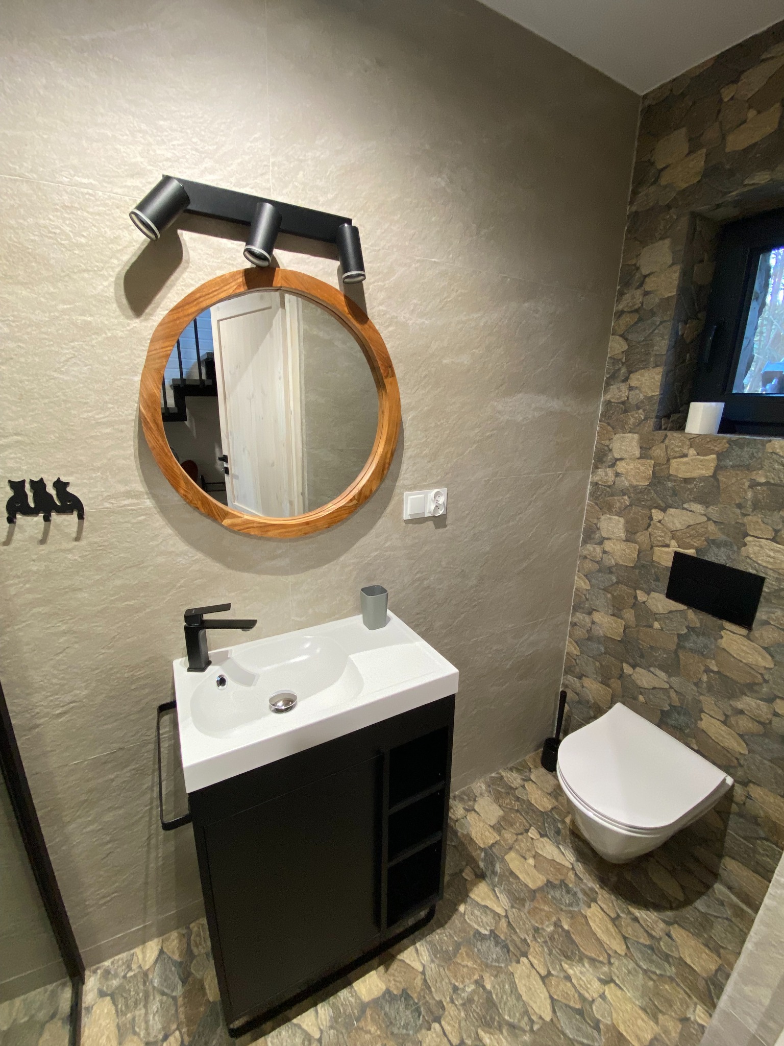 Nowoczesna łazienka z okrągłym lustrem w drewnianej ramie, czarną armaturą i kamienną mozaiką na ścianach i podłodze. Minimalistyczny design z akcentami naturalnych materiałów.