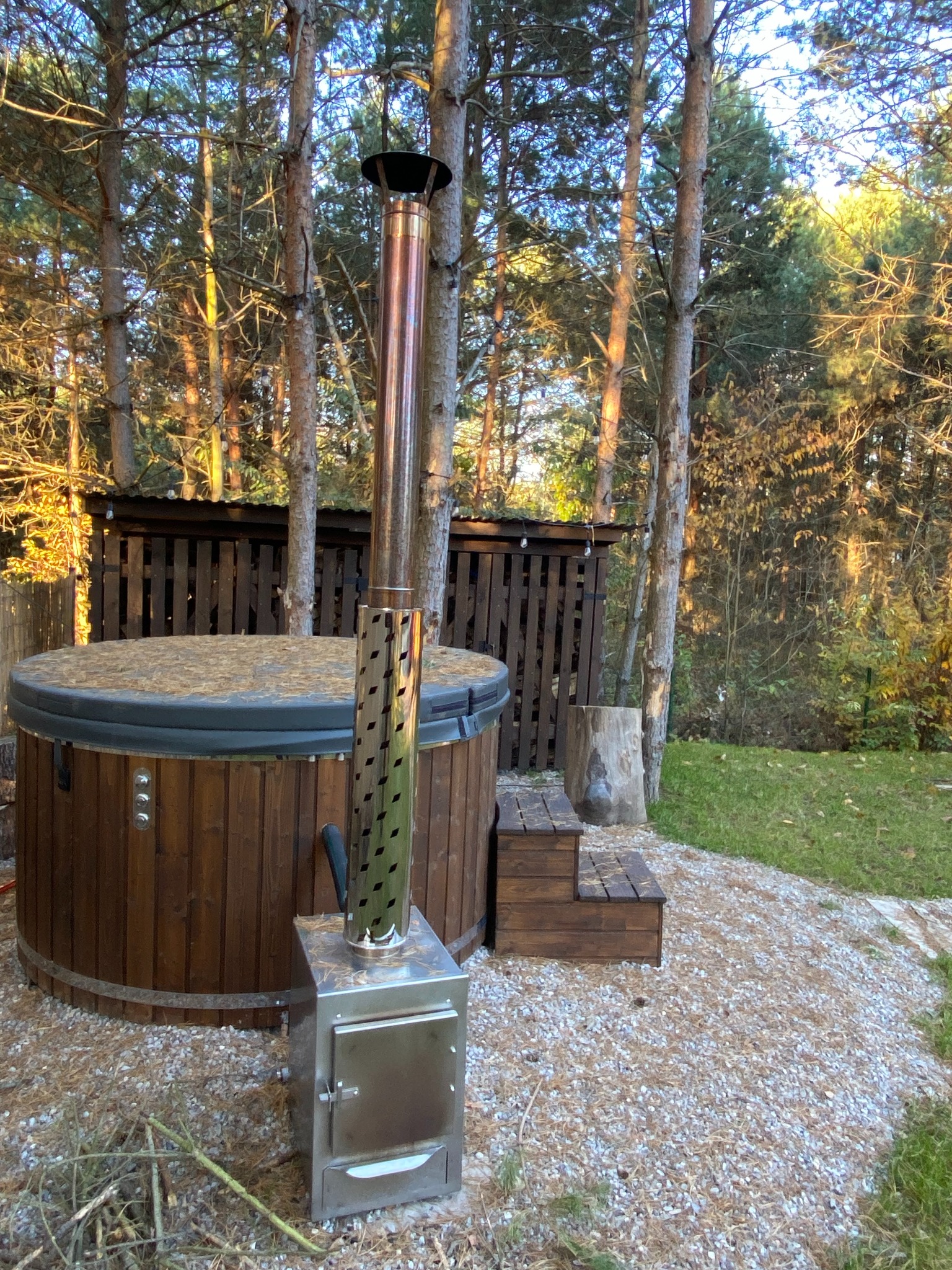 Drewniana beczka z piecem na drewno i kominem na zewnątrz, otoczona drzewami i żwirowym podłożem. Widoczne drewniane schodki i zadaszenie na drewno w tle.