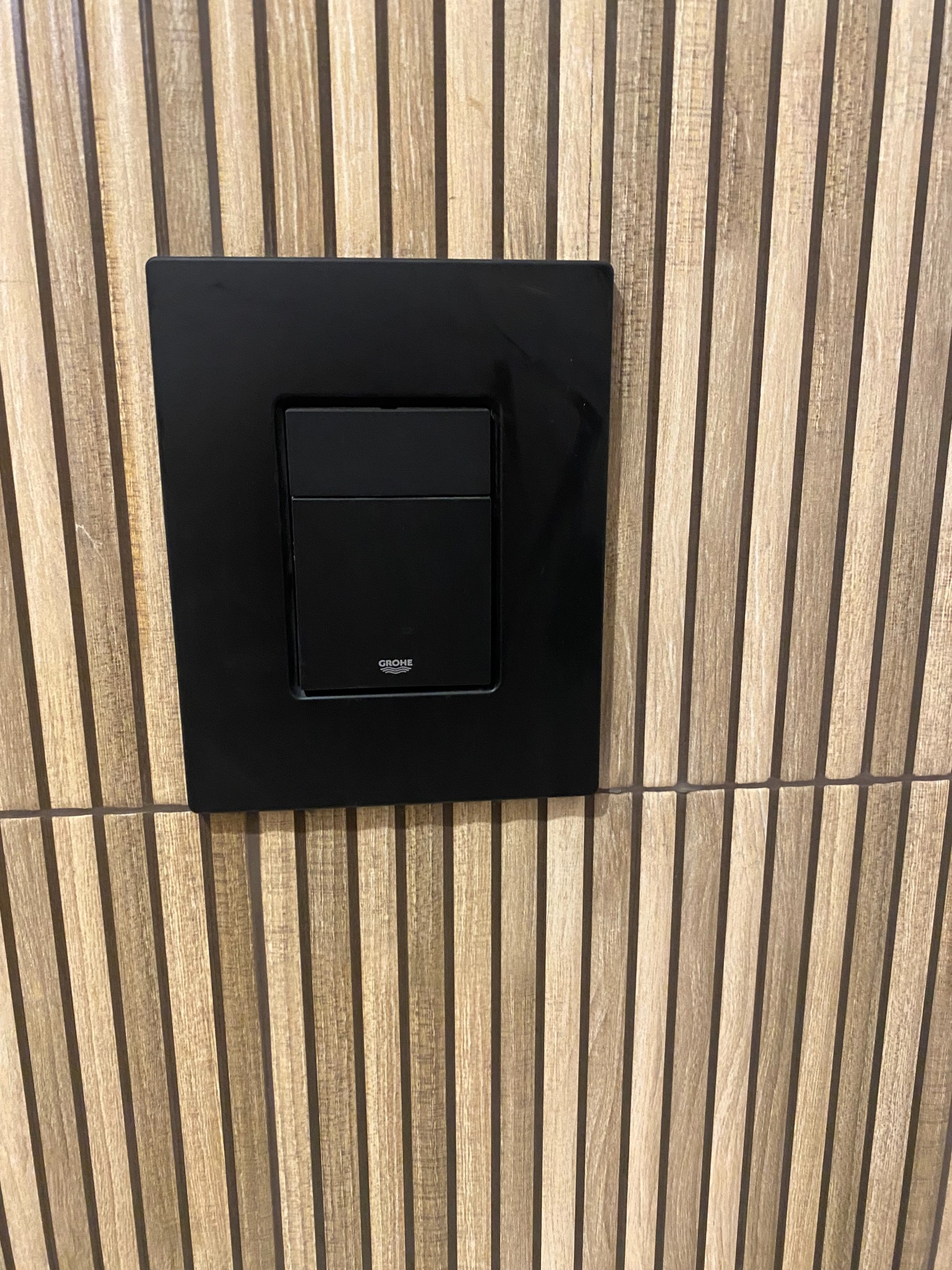Czarny panel spłukujący Grohe na ścianie z drewnopodobnych paneli. Minimalistyczny design, nowoczesne wykończenie łazienki, detale armatury.