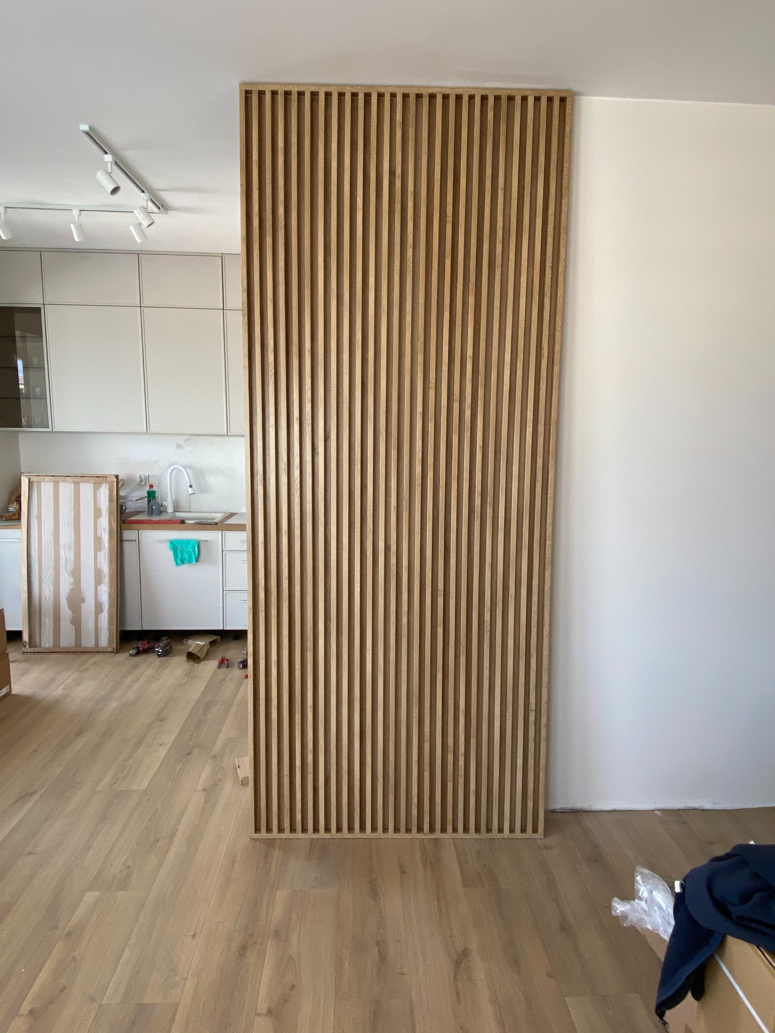 Drewniana ścianka działowa z listew w jasnym wnętrzu, widoczna kuchnia w tle. Element dekoracyjny oddzielający przestrzeń, surowe drewno, minimalistyczny design.