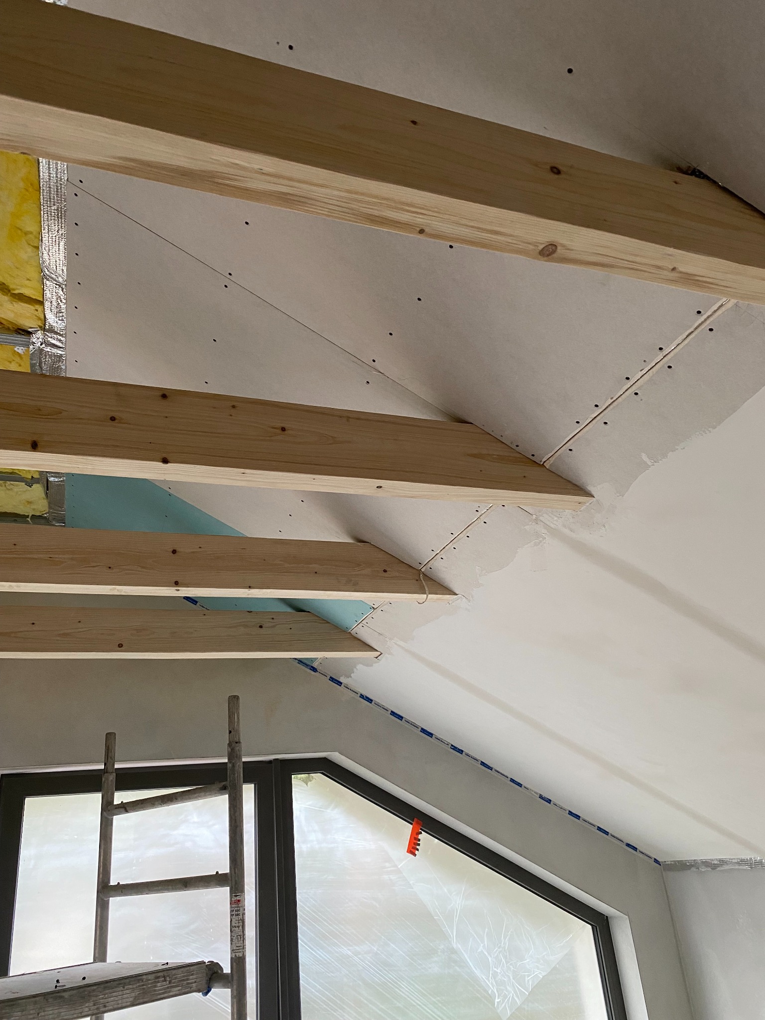 Skośny sufit poddasza w trakcie wykańczania: drewniane belki, płyty gipsowe, izolacja, okno z folią ochronną, drabina. Widok z dołu, etap prac wykończeniowych.