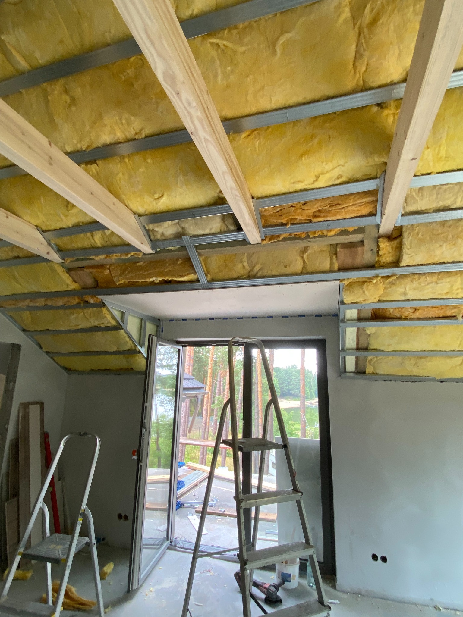 Ocieplenie poddasza wełną mineralną, widoczne metalowe profile i drewniane belki. W tle okno balkonowe i drabina. Prace wykończeniowe w trakcie realizacji.