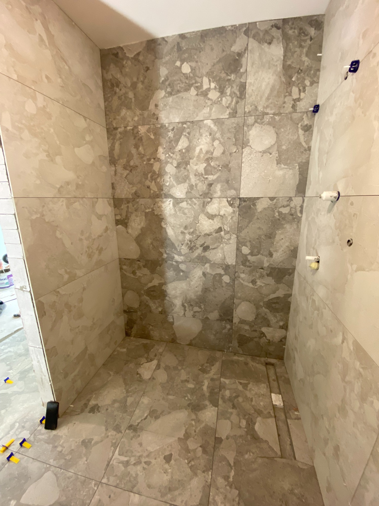 Wykończona kabina prysznicowa z szarymi płytkami imitującymi marmur. Widoczne niebieskie krzyżyki dystansowe i odpływ liniowy w podłodze. Minimalistyczny design.