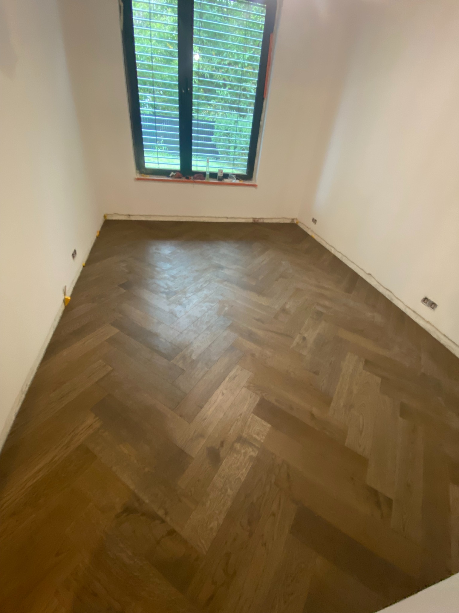 Ukończona podłoga z parkietu ułożonego w jodełkę w pustym pokoju z oknem. Widoczne listwy przypodłogowe i żółte kliny dystansowe.