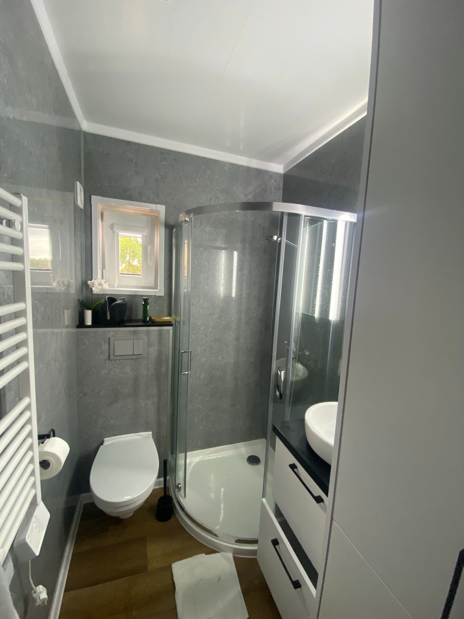 Nowoczesna łazienka w szarościach z kabiną prysznicową, umywalką na szafce i toaletą. Na ścianie grzejnik drabinkowy i okno z dekoracjami. Drewnopodobna podłoga.