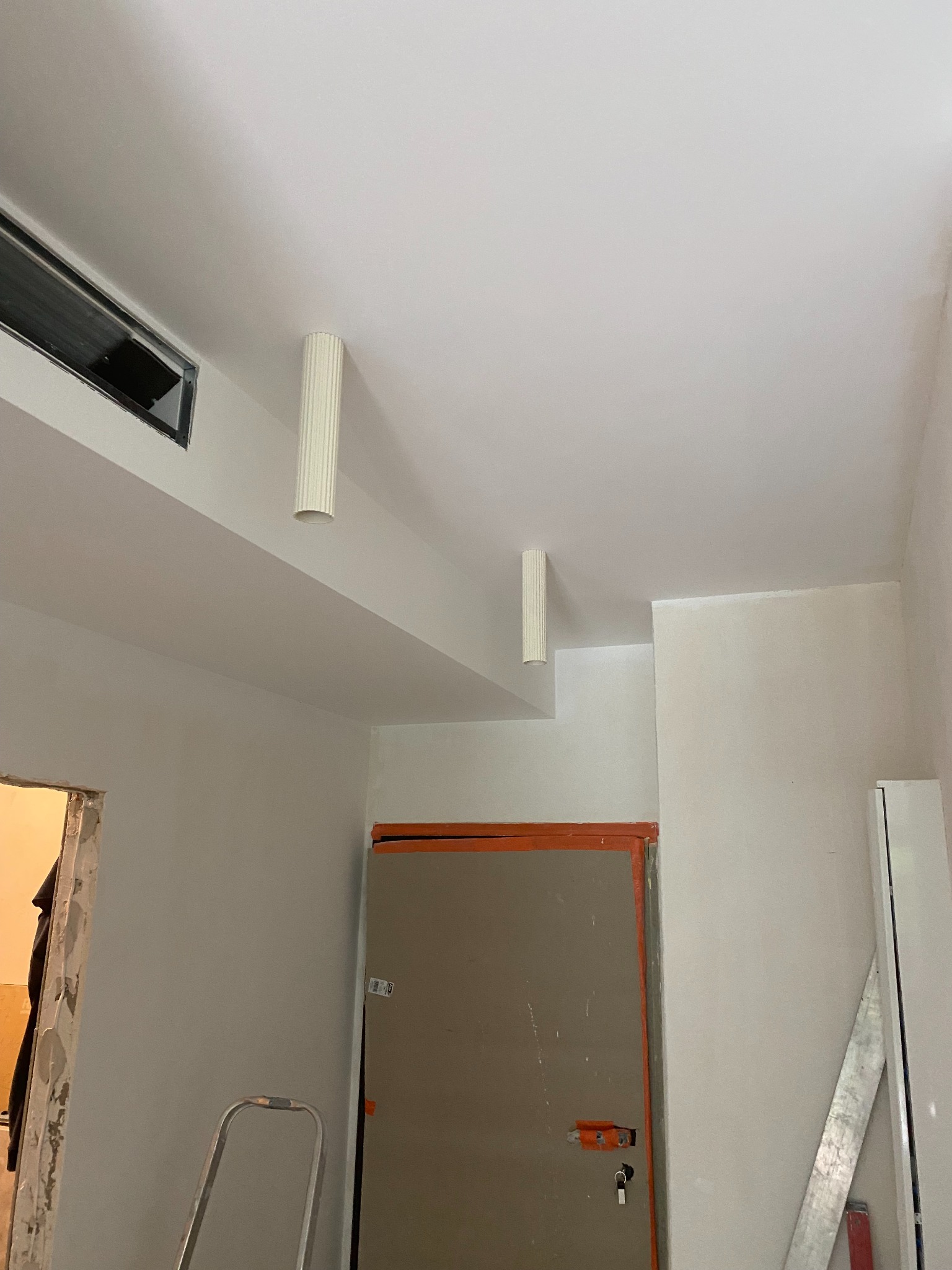 Wnętrze w trakcie remontu: pomalowany sufit z dwoma cylindrycznymi lampami, drzwi z pomarańczową ramą, drabina i listwa przypodłogowa. Widoczne ślady prac remontowych.