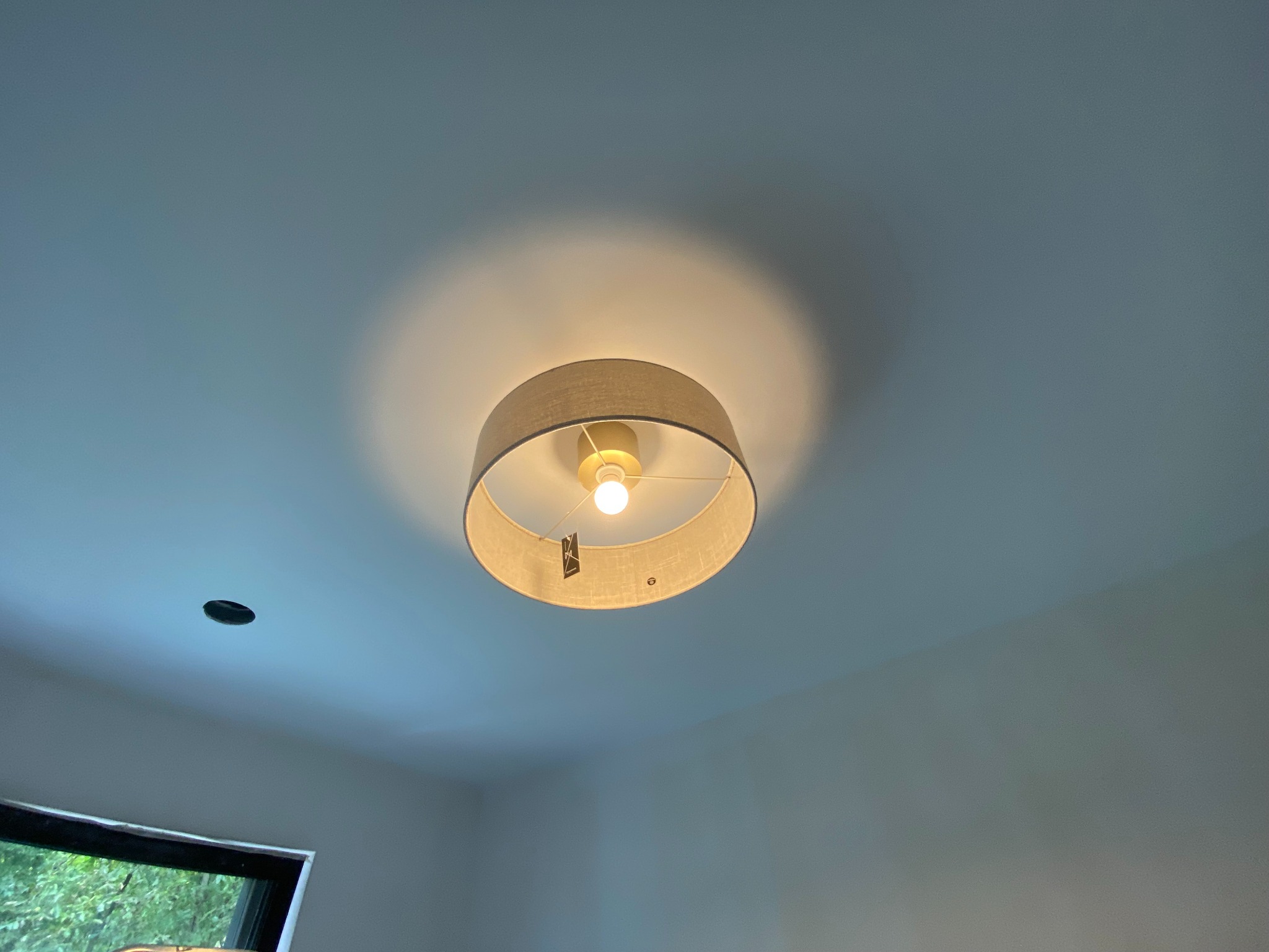 Zamontowana okrągła lampa sufitowa z abażurem z tkaniny, oświetlająca biały sufit. Widoczny otwór po starej lampie i fragment okna w dolnym rogu.
