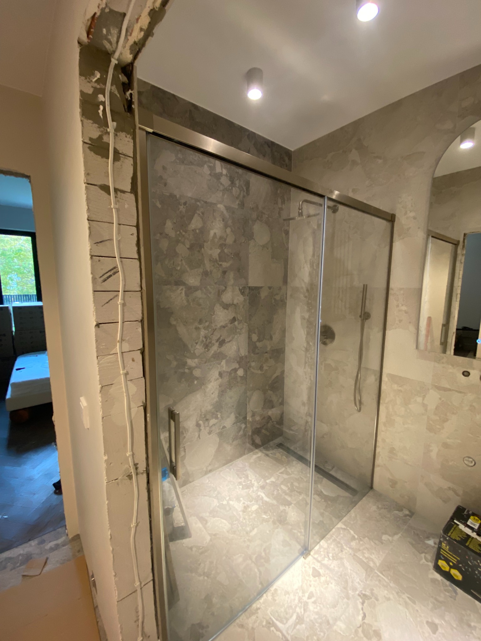 Szklana kabina prysznicowa z szarymi płytkami imitującymi marmur, widoczna w trakcie montażu. Otwarta przestrzeń z widocznymi elementami instalacji elektrycznej.