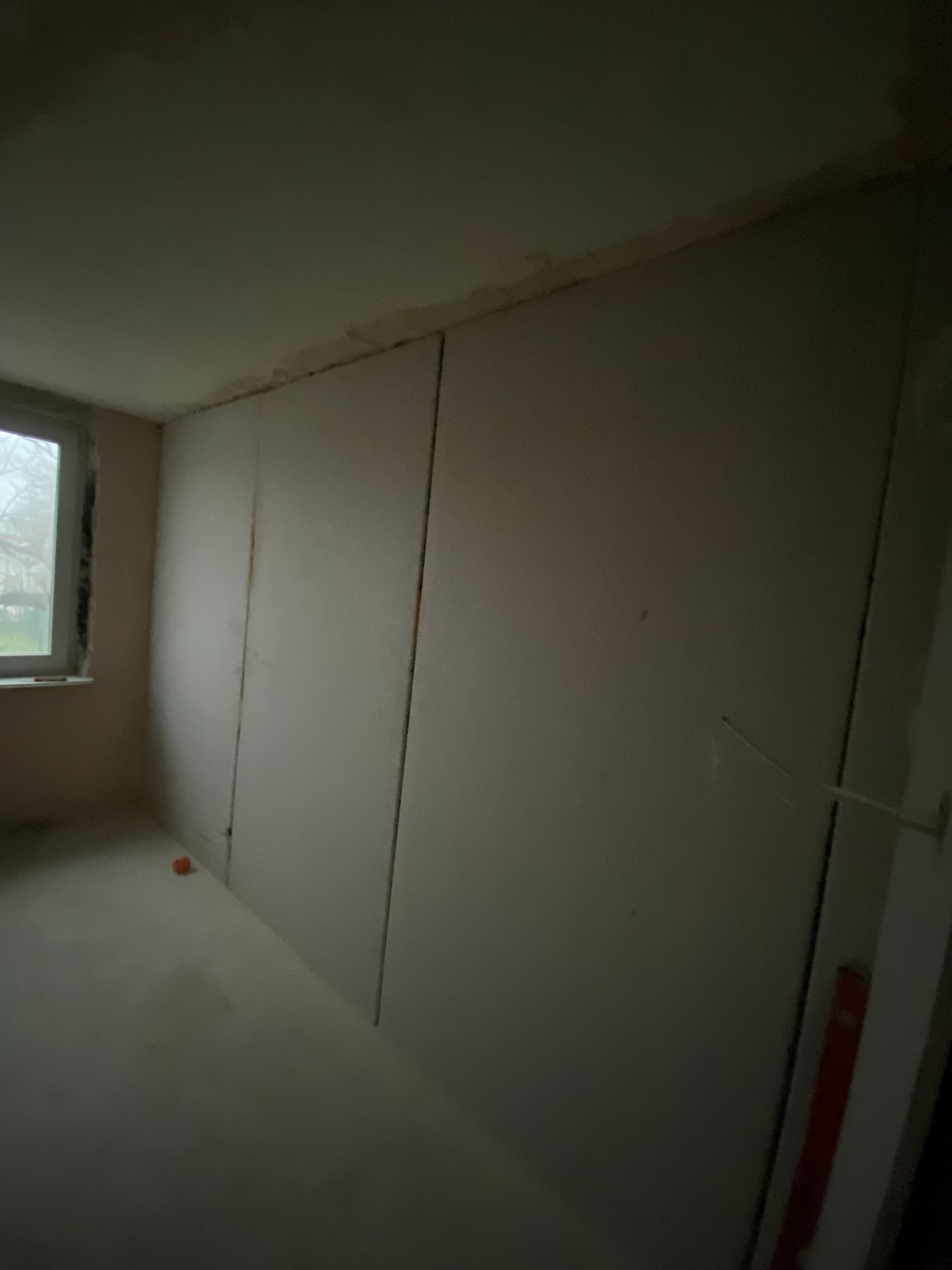 Wnętrze w trakcie prac wykończeniowych: ściany pokryte płytami gipsowo-kartonowymi, widoczne łączenia. Okno z widokiem na zewnątrz. Na podłodze leży pomarańczowy klocek.