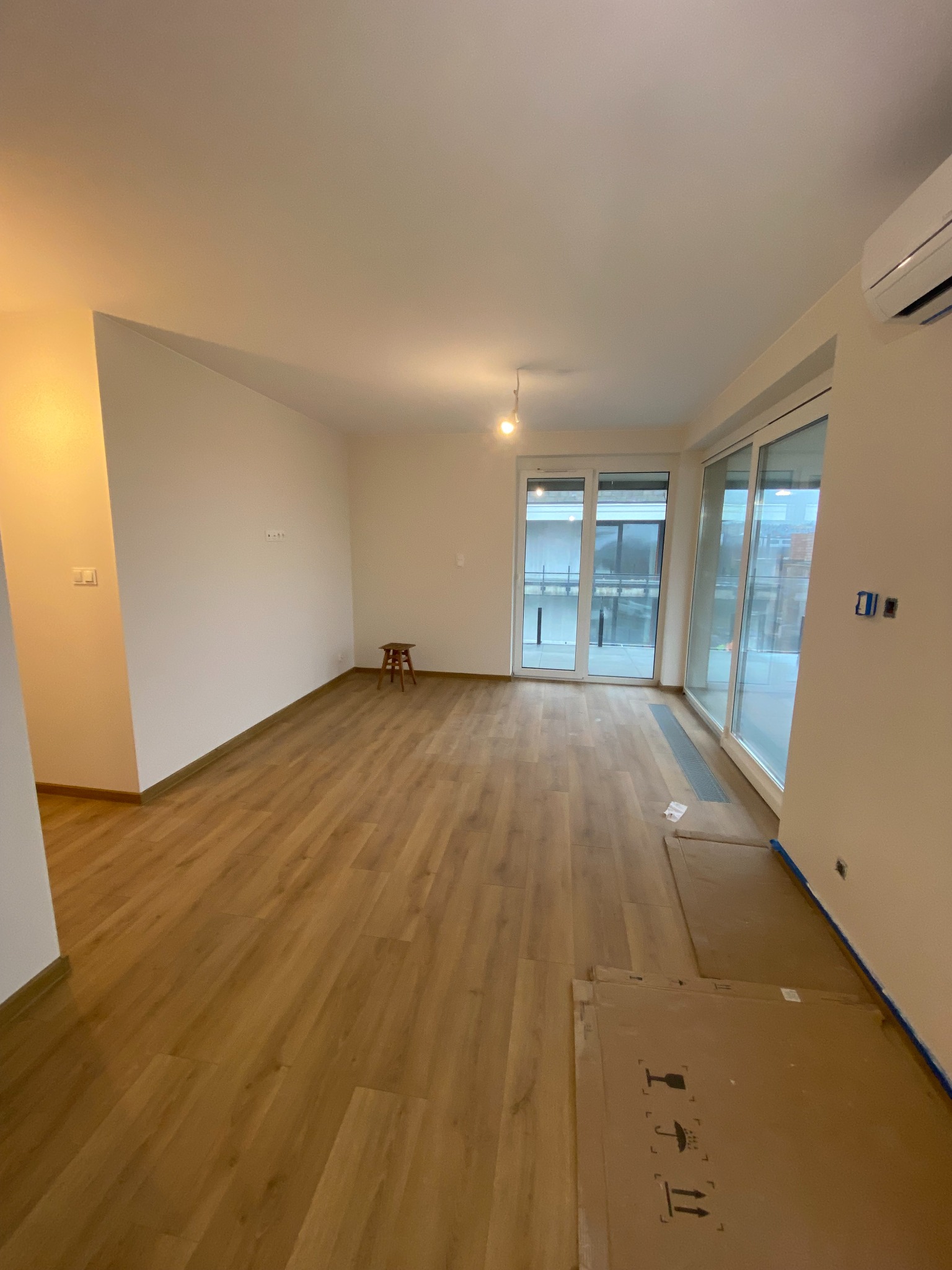 Jasne, przestronne wnętrze po remoncie z drewnianą podłogą i dużymi oknami balkonowymi. Minimalistyczny wystrój z małym stołkiem i kartonami na podłodze.