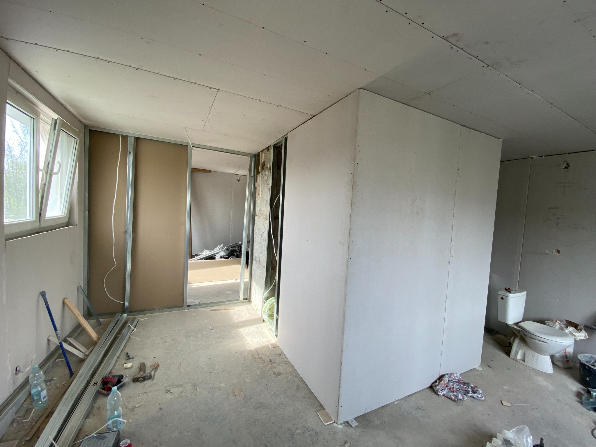 Wnętrze w trakcie prac wykończeniowych: ściany działowe z płyt gipsowo-kartonowych, widoczne profile metalowe, narzędzia i materiały budowlane na podłodze. Częściowo wykończona łazienka.