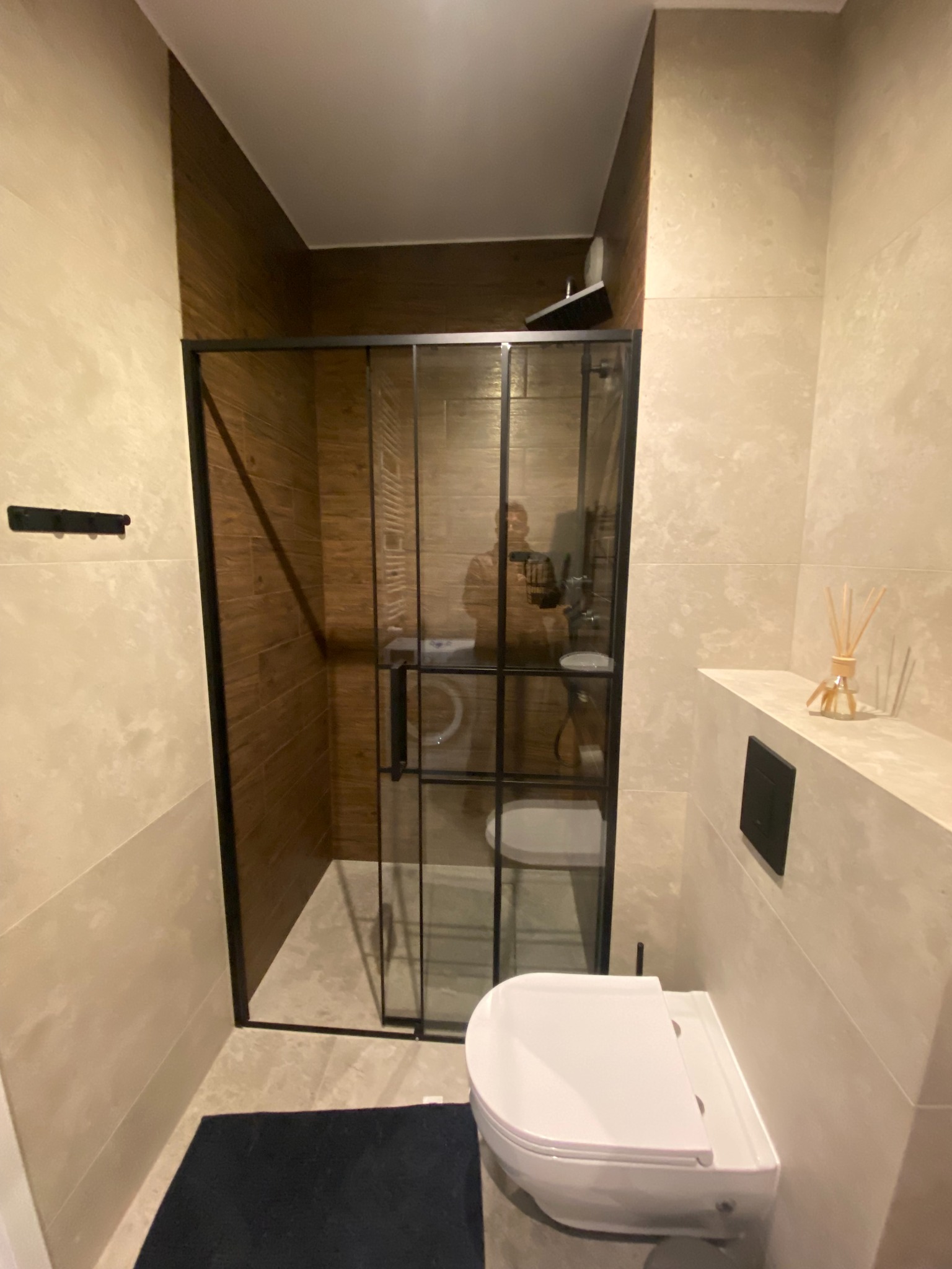 Nowoczesna łazienka z kabiną prysznicową typu walk-in z czarnymi ramami, beżowymi płytkami i drewnianą ścianą wewnątrz kabiny. Widoczna toaleta i dywanik.