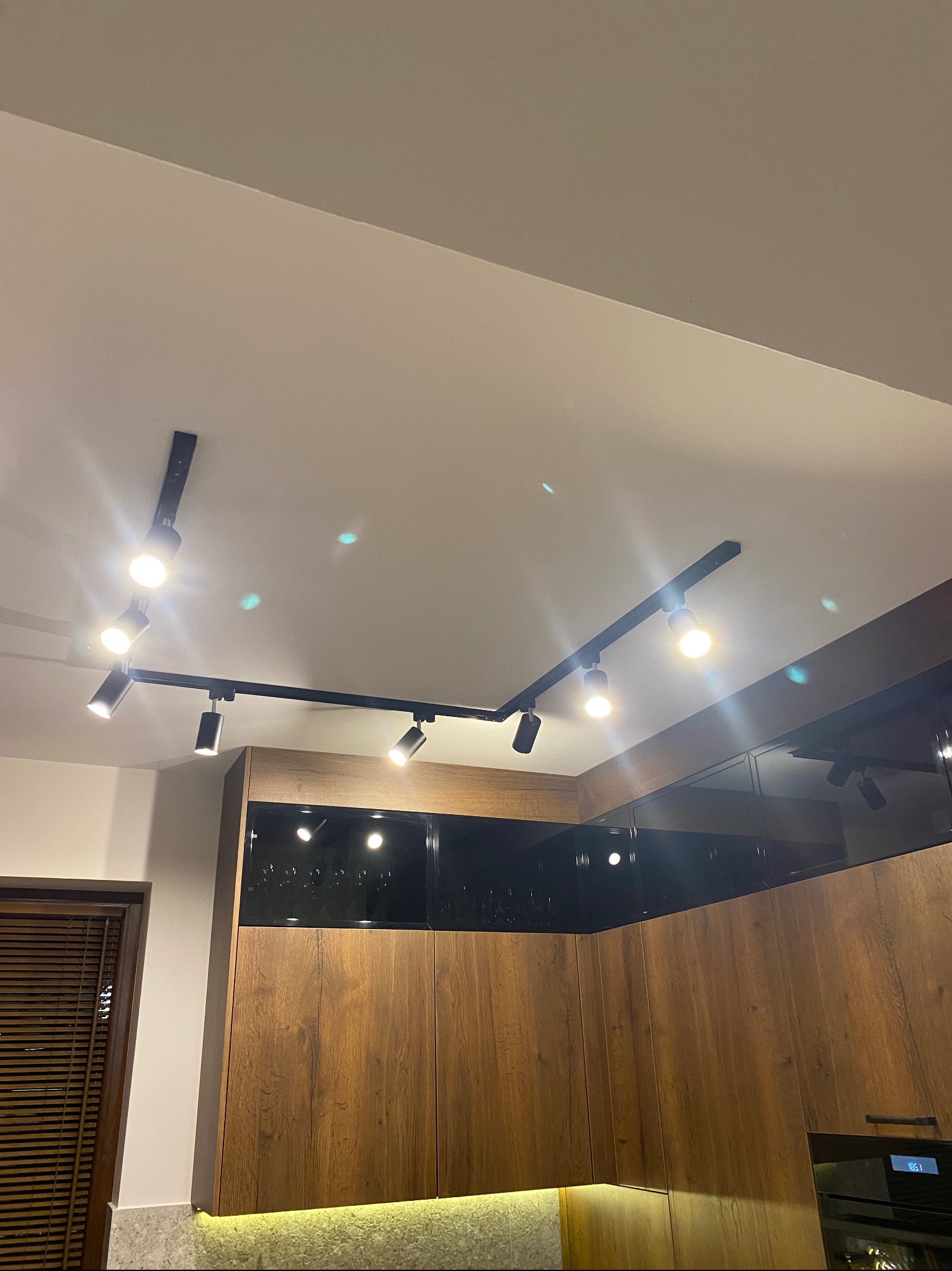 Nowoczesna kuchnia z drewnianymi szafkami, czarnymi frontami i oświetleniem szynowym. Widok z dołu na sufit z lampami punktowymi.