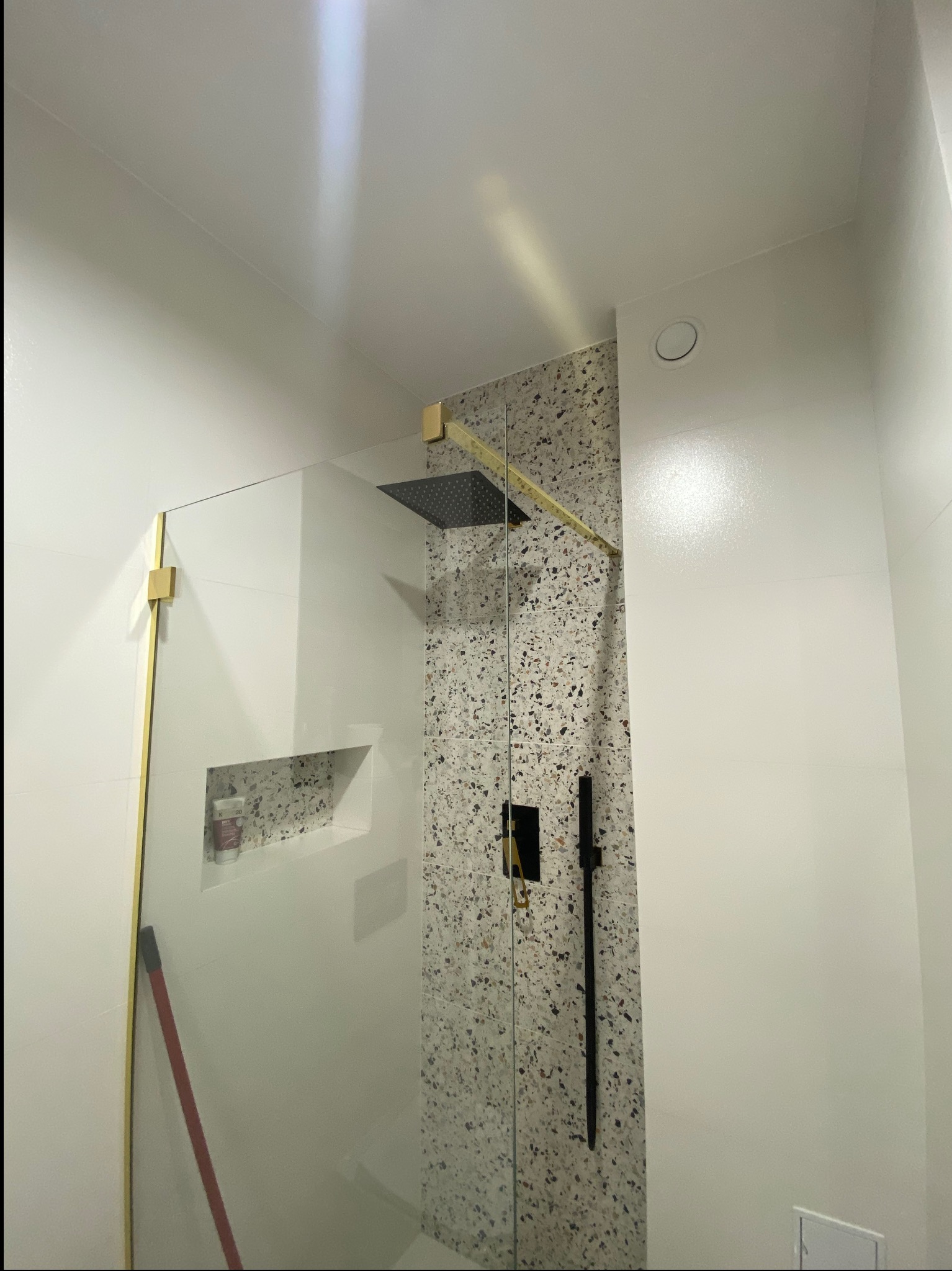 Nowoczesna kabina prysznicowa z deszczownicą, wykończona płytkami lastryko i złotymi detalami, w jasnej łazience. Widoczna wnęka z kosmetykami.