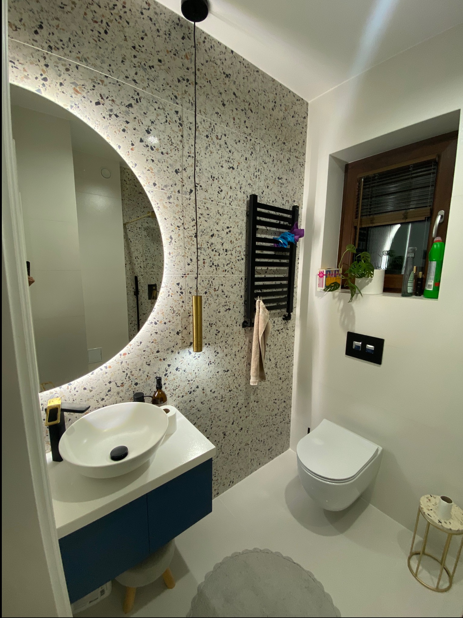 Nowoczesna łazienka z okrągłym lustrem LED, umywalką nablatową i płytkami lastryko. Grzejnik drabinkowy i toaleta pod oknem dopełniają minimalistyczny wystrój.