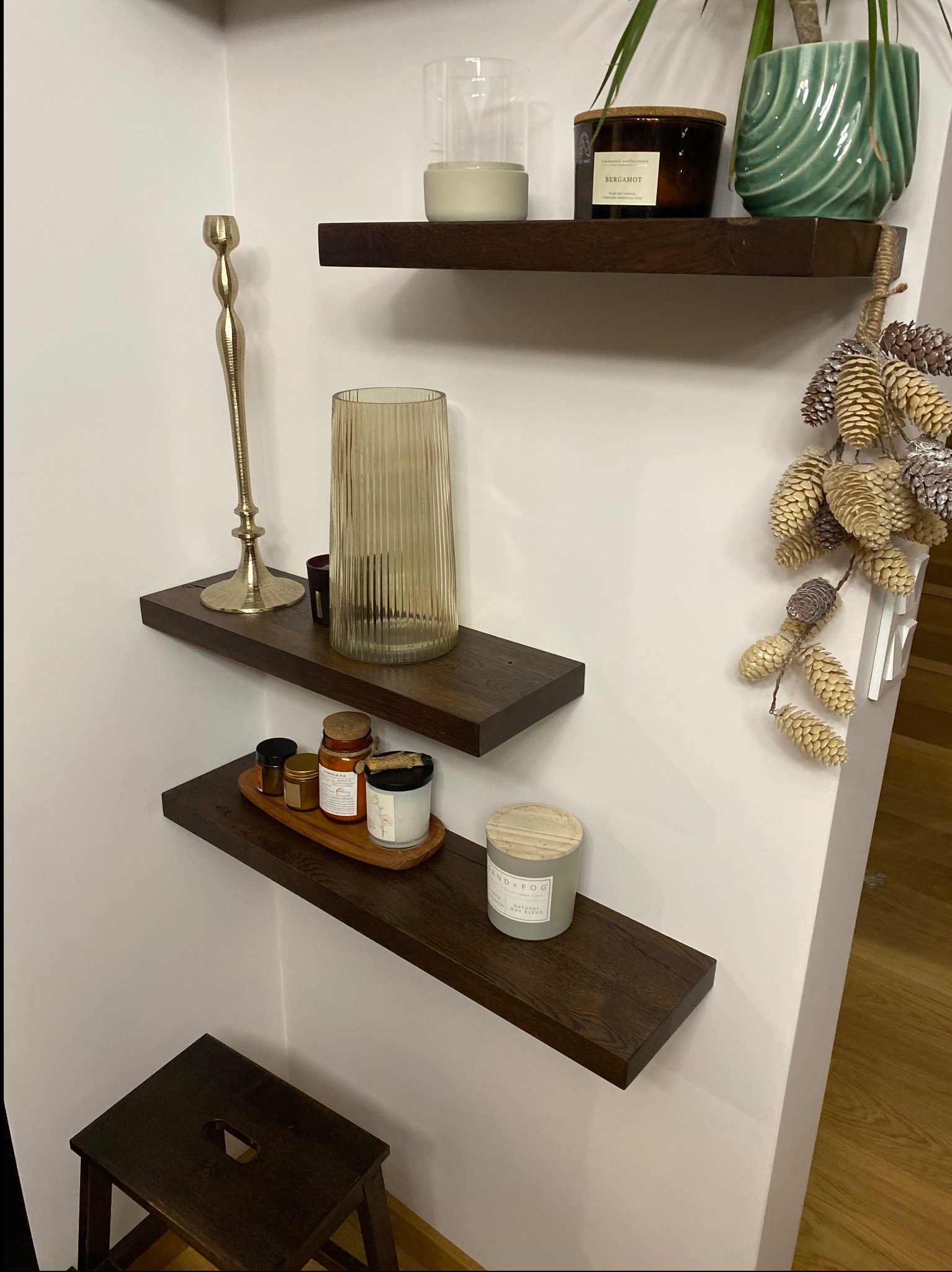 Trzy ciemne, drewniane półki na białej ścianie, udekorowane świecami, wazonami i roślinami. Na dole mały, drewniany stołek. Dekoracja wnętrza w ciepłych barwach.