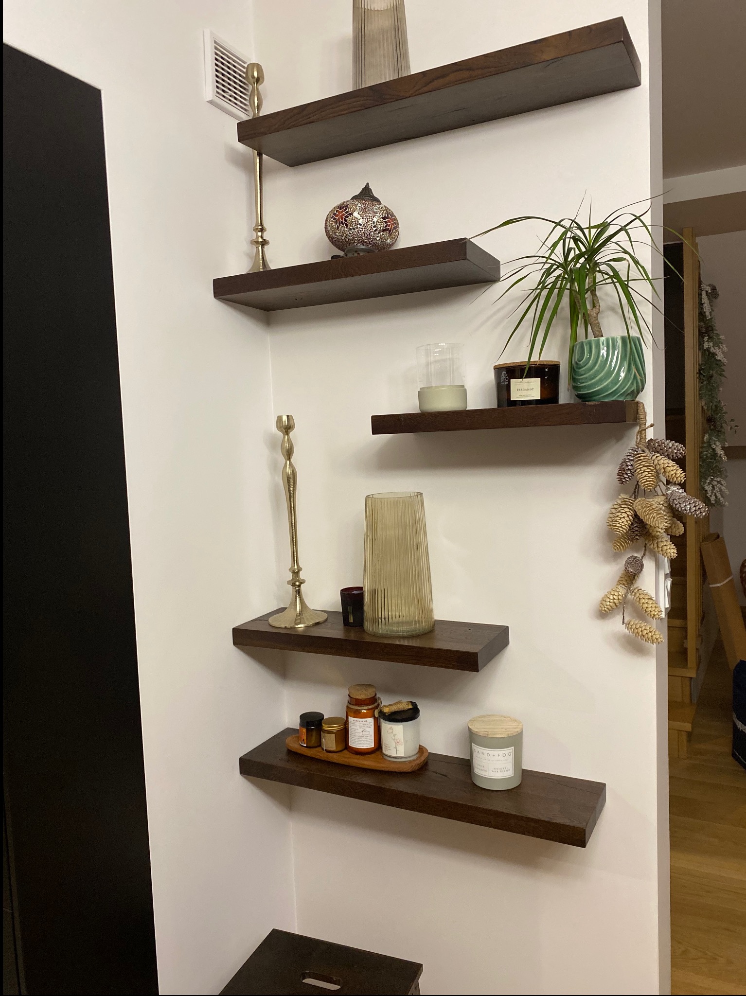 Ciemne, drewniane półki zamontowane na białej ścianie, na których stoją dekoracje: świece, wazon, roślina doniczkowa i ozdobna lampka. Widok z dołu, oświetlenie naturalne.