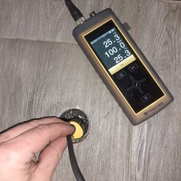 MM - OSUSZANIE MICHAŁ MIELKAU - Pomiar wilgotności posadzki za pomocą wilgotnościomierza Trotec. Sonda w otworze, wyświetlacz pokazuje 100% wilgotności. Płytki imitujące drewno w tle.