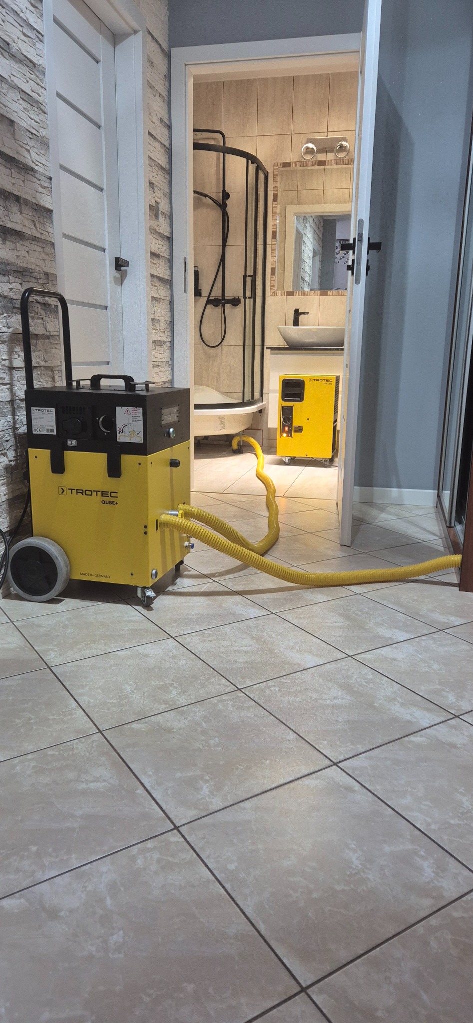 Żółty osuszacz podposadzkowy Trotec QUUBE+ z podłączonymi wężami w łazience z kabiną prysznicową. Widoczny drugi osuszacz.