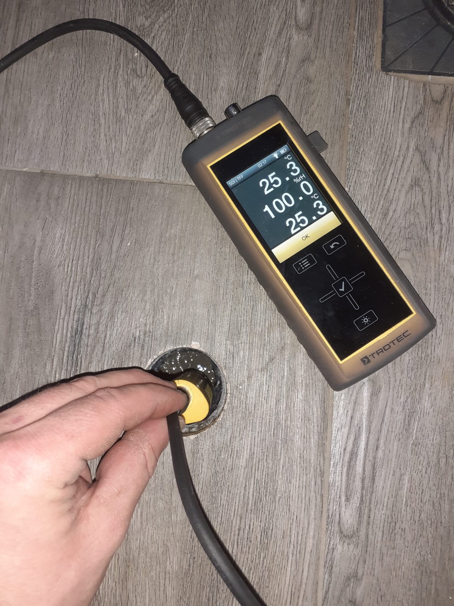 Pomiar wilgotności posadzki za pomocą wilgotnościomierza Trotec. Sonda w otworze, wyświetlacz pokazuje 100% wilgotności. Płytki imitujące drewno w tle.